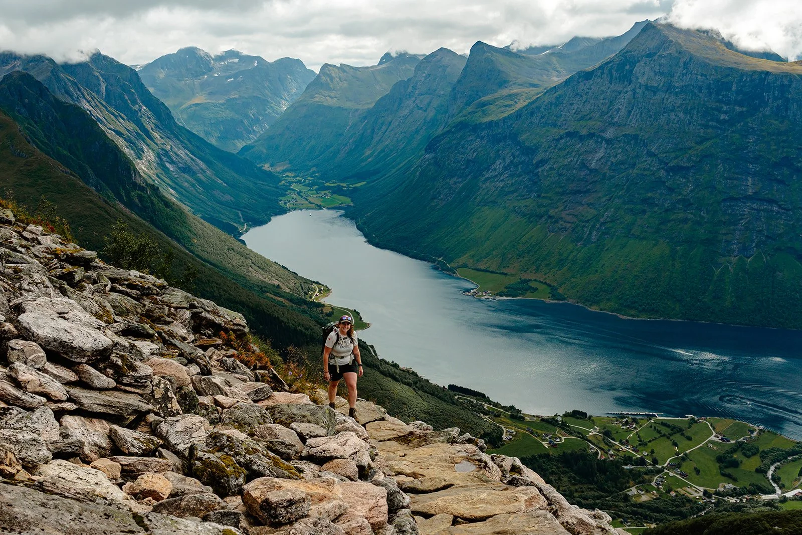A woman hiking on rocky terrain overlooking a fjord surrounded by steep, green mountains under a cloudy sky in Norwegian fjords.