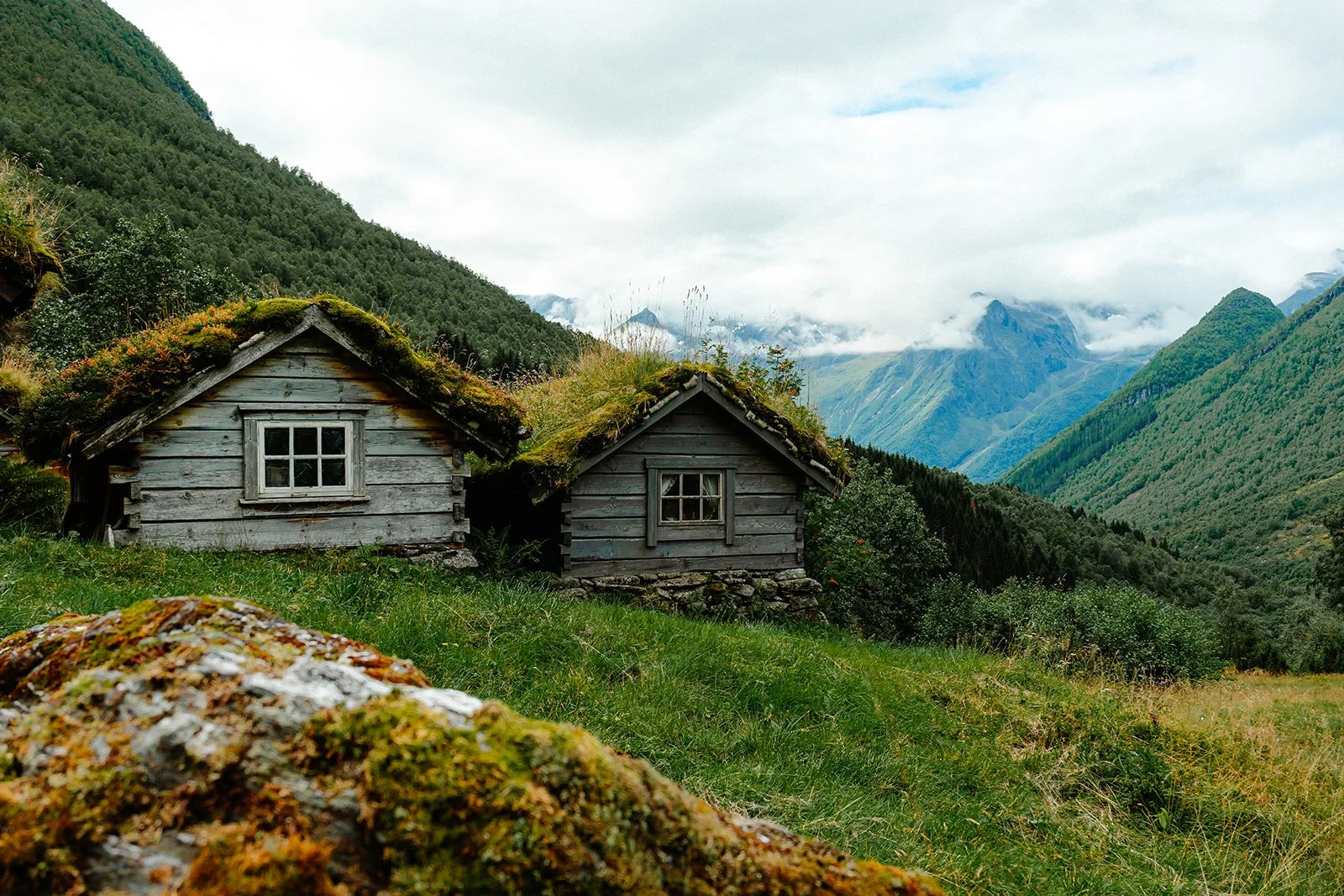 Two small wooden cabins with moss-covered roofs sit on a grassy hillside surrounded by lush mountains and clouds in the background.