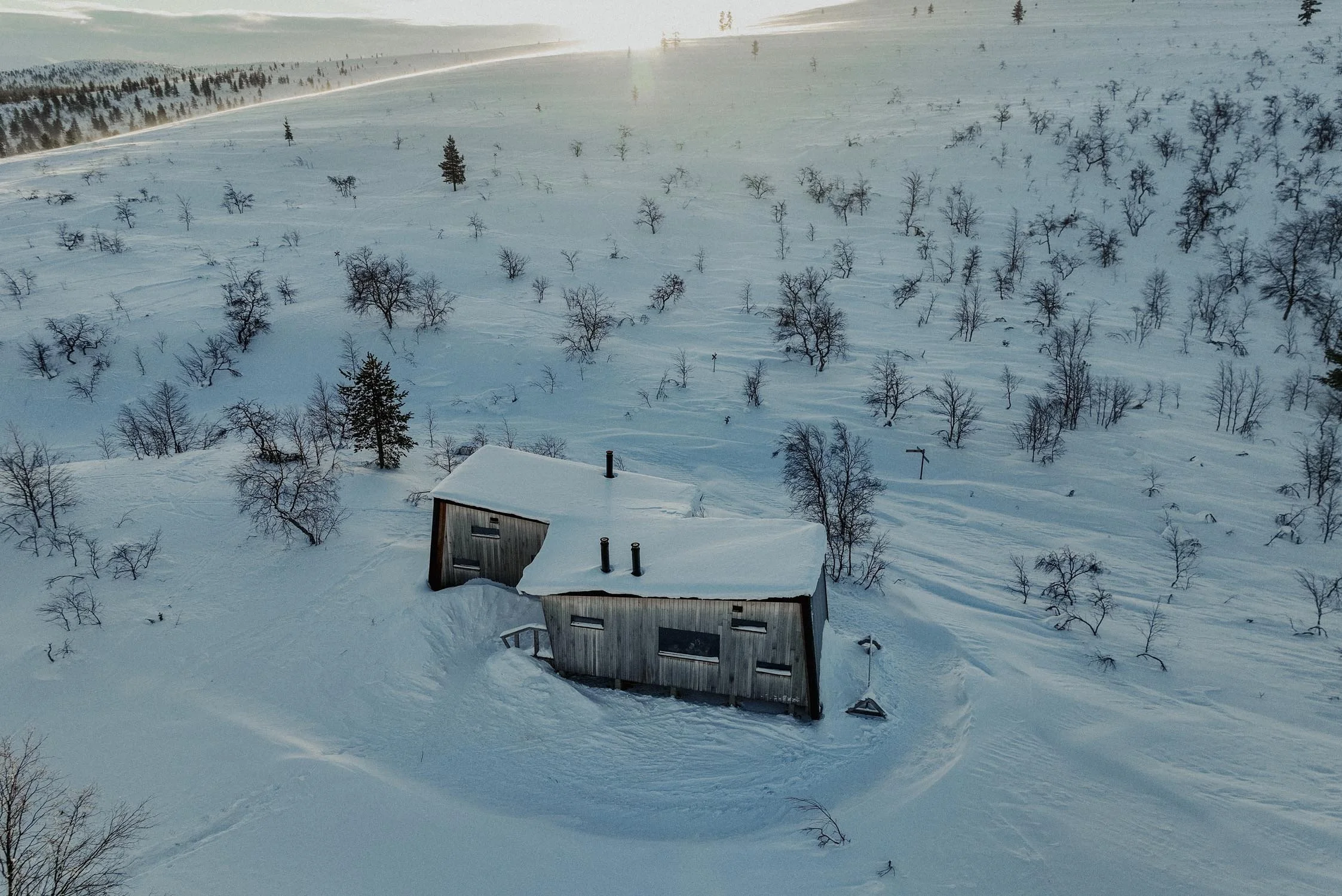 A snow-covered landscape with numerous small, leafless trees scattered across a hilly area. In the foreground, there is a small house with a snow-covered roof and four chimney pipes.