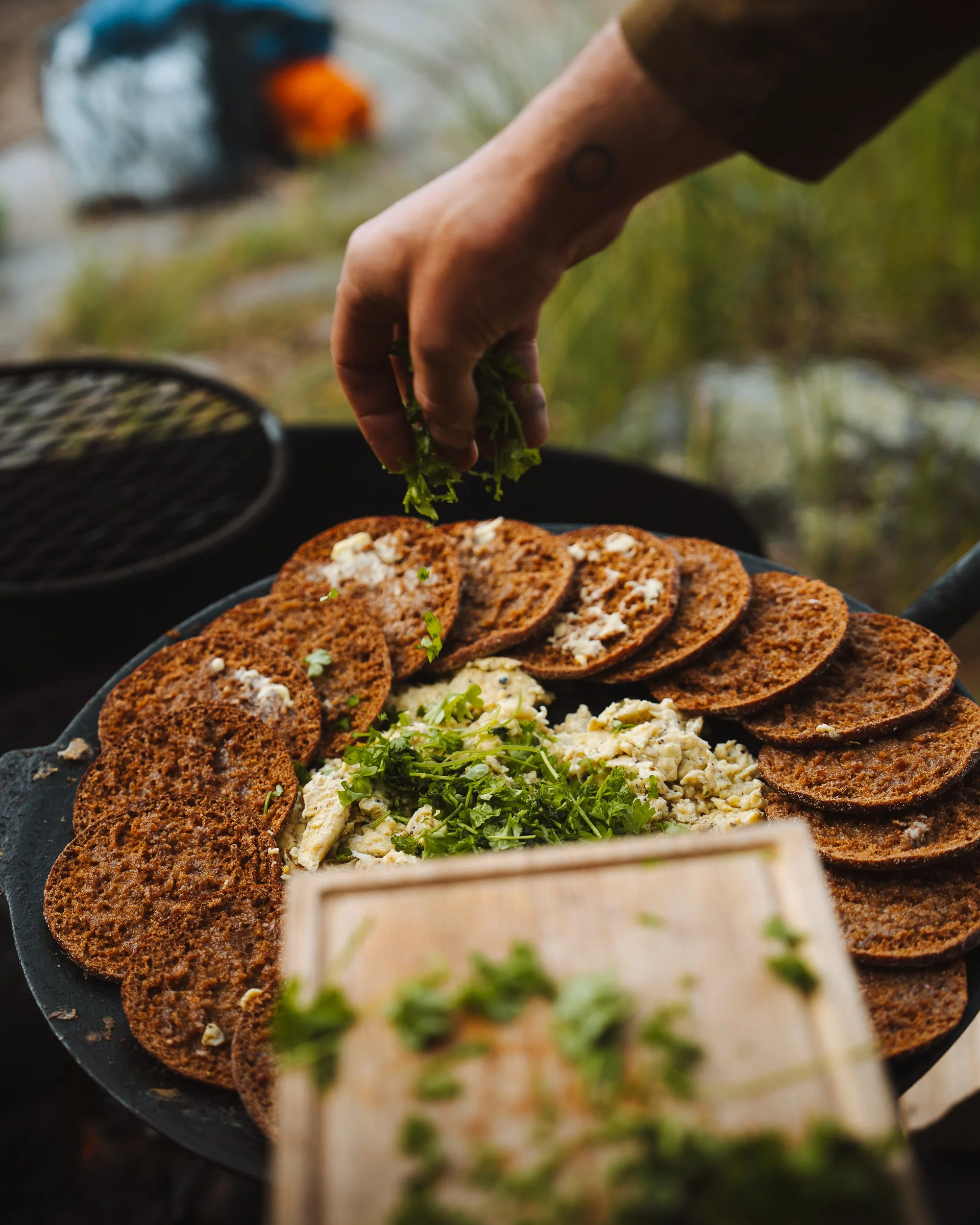 Close-up of a person's hand placing fresh green herbs on a plate of food, with slices of dark brown bread and a creamy dish topped with herbs, outdoors near a lake with fishing boats in the background.