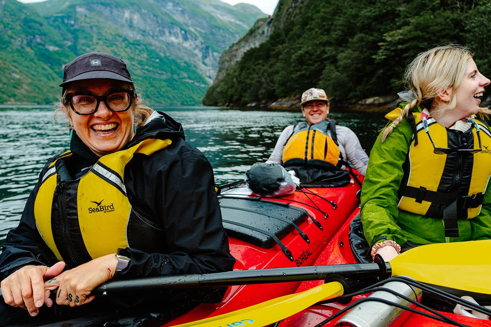 Three people on a red kayak enjoying a scenic view of a river surrounded by green mountains, all wearing life jackets and outdoor gear.