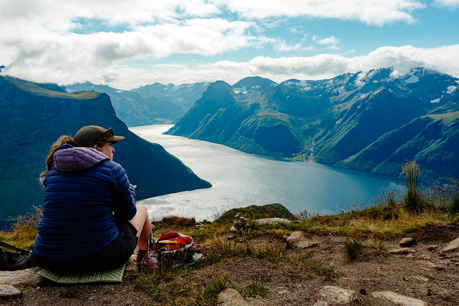 A woman sitting on a grassy hill overlooking a fjord with mountains in the background, wearing outdoor clothing and a cap, with backpacks nearby.