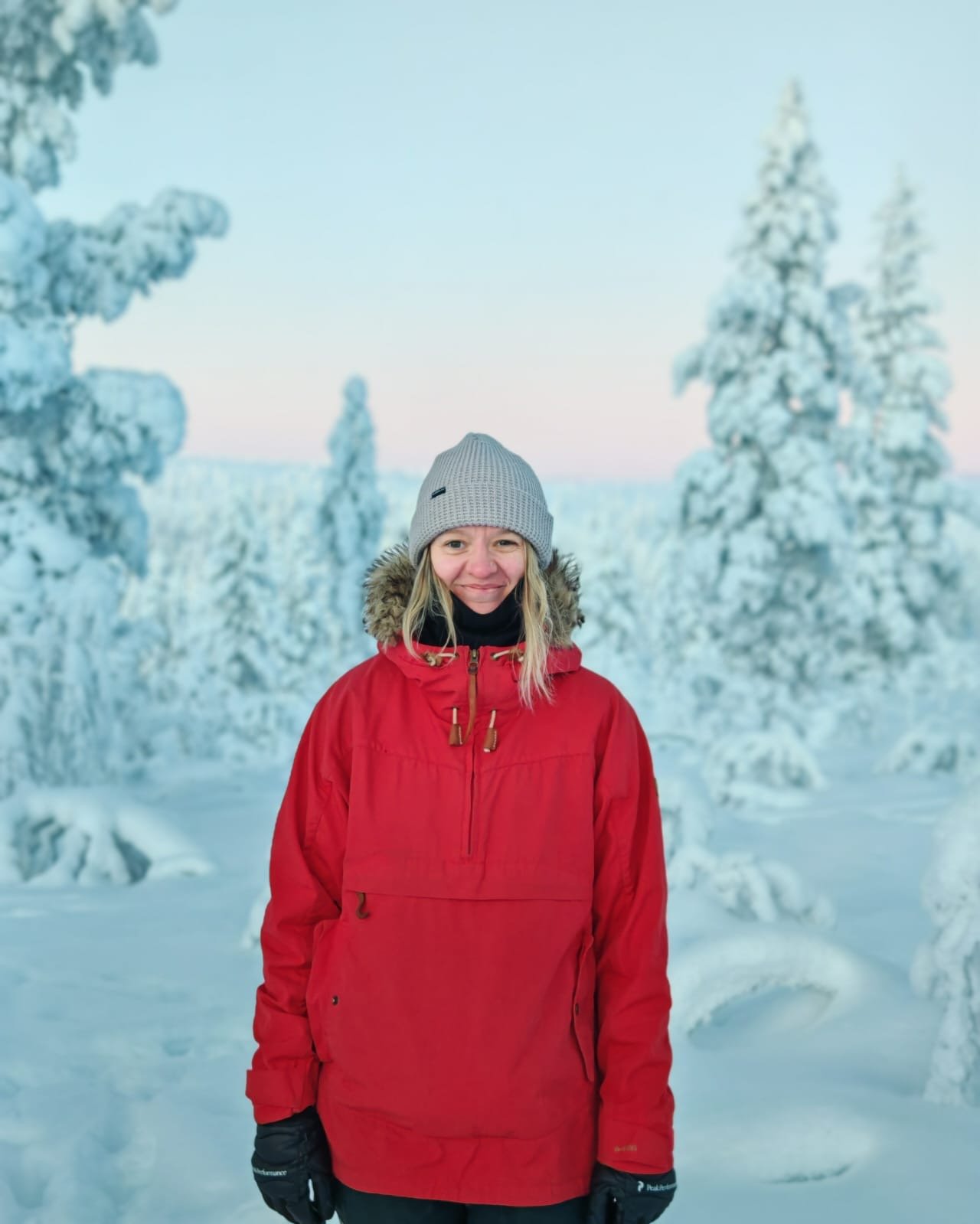 A woman in a red winter jacket, gray knit hat, and black gloves standing outdoors in a snow-covered landscape with snow-laden trees in the background.