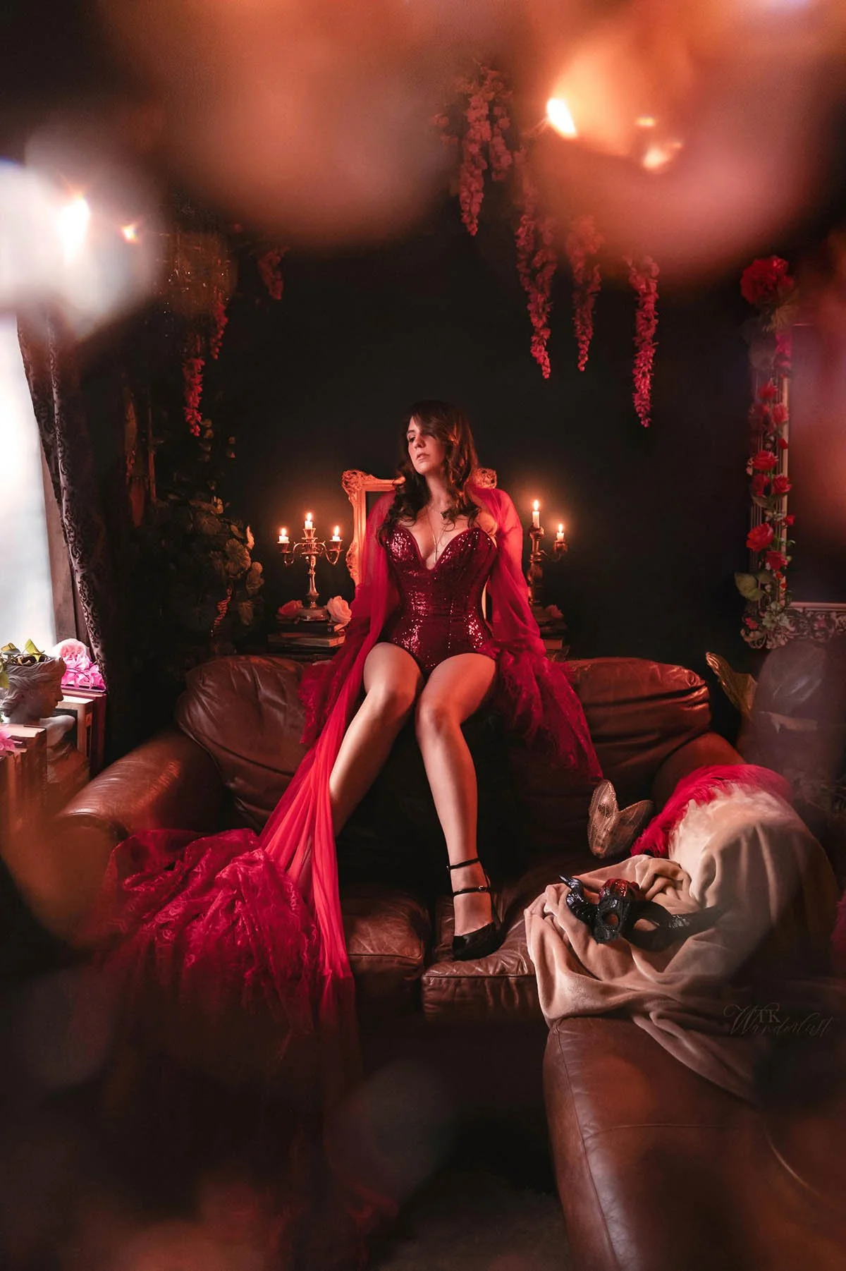 Photo of Noel Nichols, who is a petite mixed indigenous queer woman. She has wavy brown hair and is wearing a red sequined corset, a red robe and black high heels. She is sitting on the back on a dark brown leather couch.