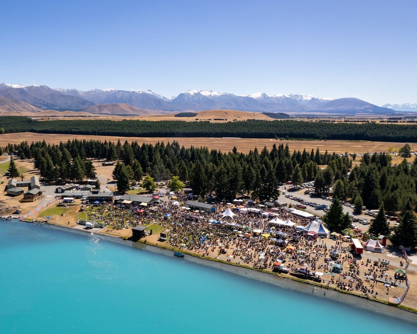 Aerial view of a lakeside festival with crowds, marquees and mountains