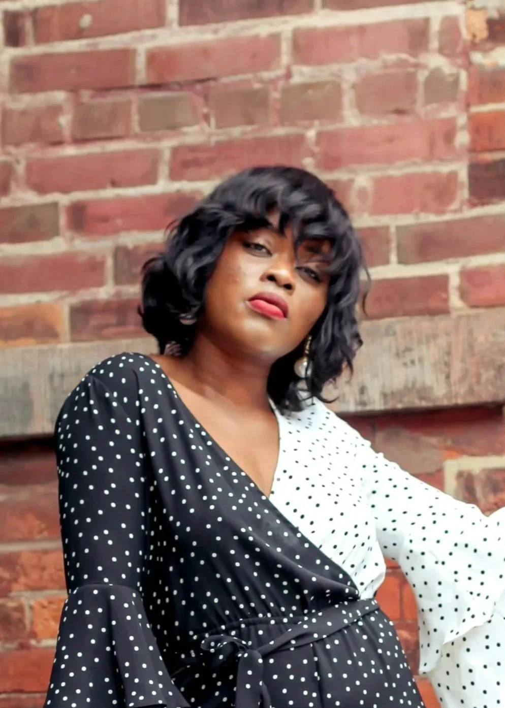 A woman with dark, curly hair, wearing a black and white polka dot dress with a V-neckline, standing against a brick wall.