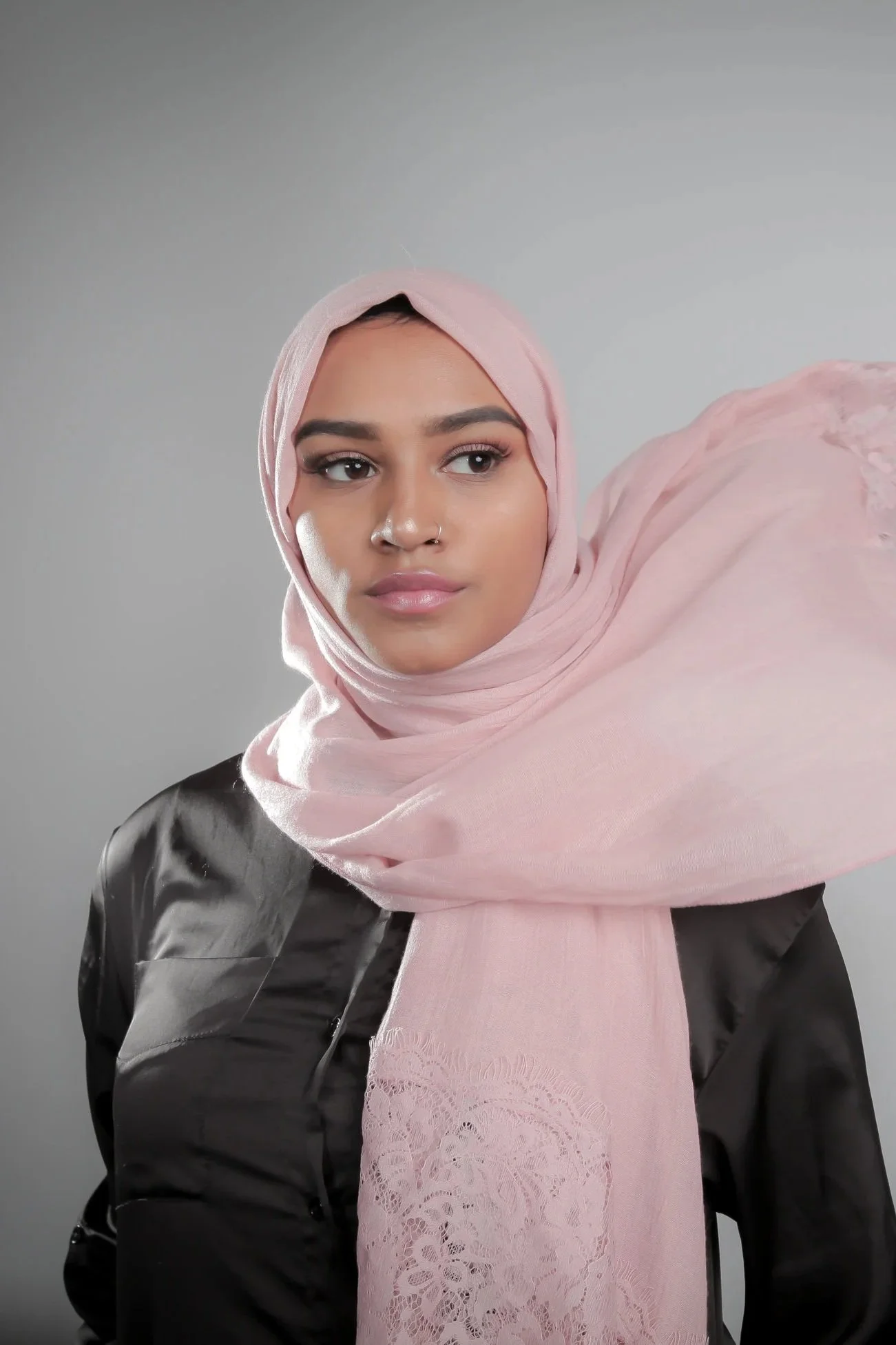 A young woman with medium skin tone wearing a pink headscarf and a black leather jacket against a plain gray background.