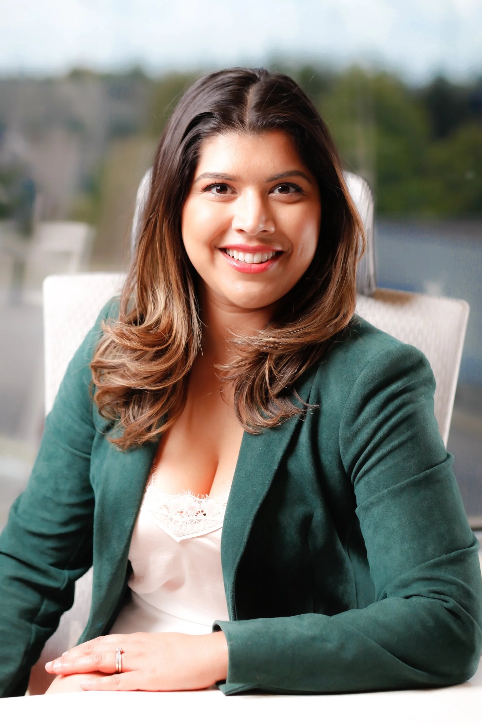 A woman with shoulder-length brown hair, smiling, sitting at a desk, wearing a green blazer and white lace top, with blurred outdoor scenery in the background.