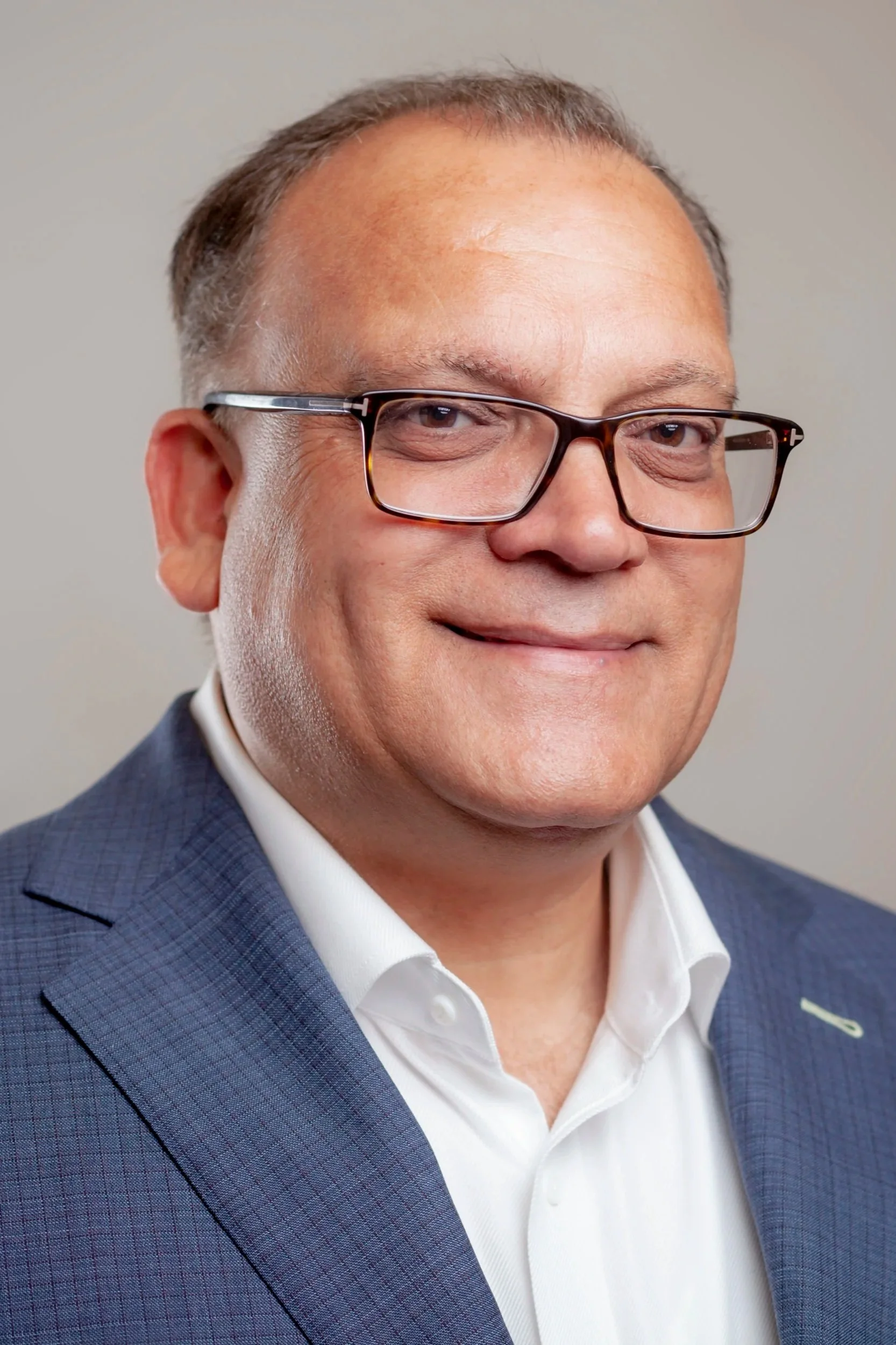 Close-up of a smiling man wearing glasses, a dark blue checked blazer, and a white shirt, against a plain light-colored background.