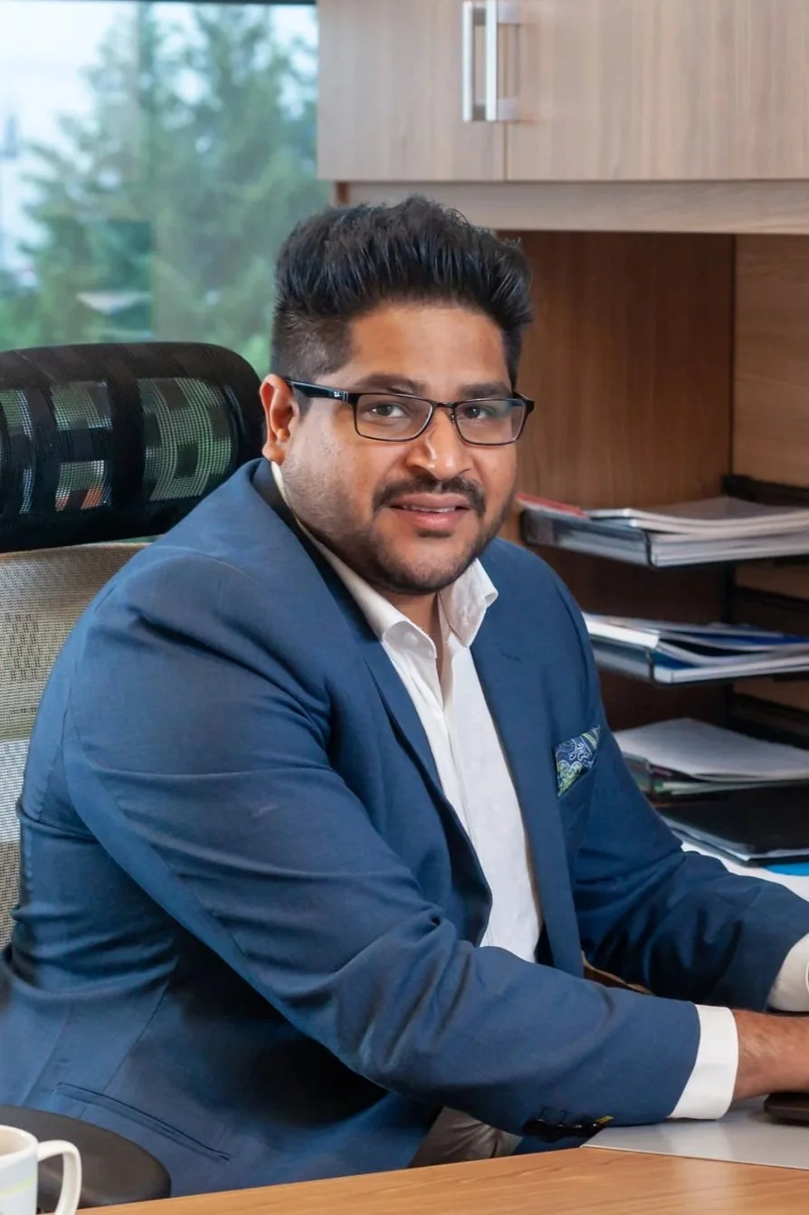 A man in a blue suit and glasses working on a laptop at a desk in an office with wooden cabinets and a large window showing a green outdoor area.