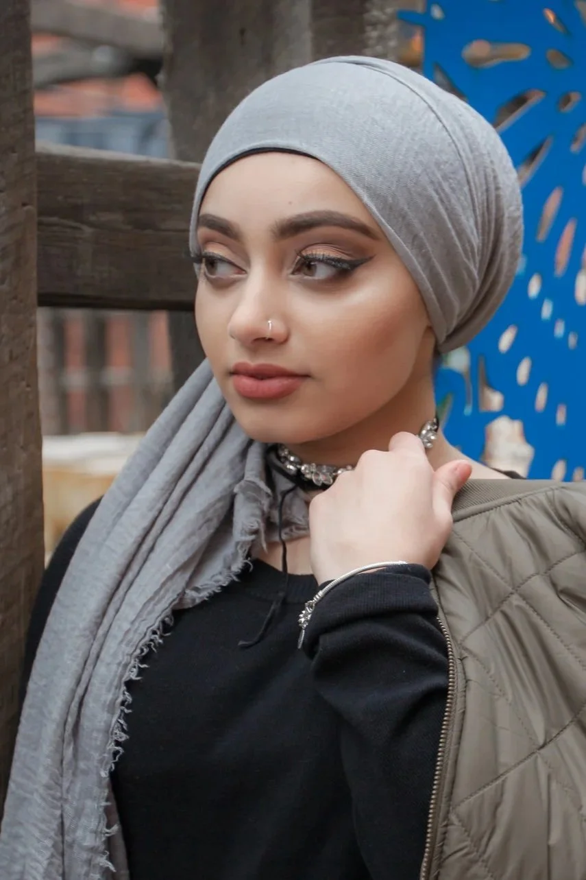 A young woman with light skin, wearing a grey hijab, black shirt, and an olive green quilted jacket, standing outdoors against a wooden fence with a blue decorative metal panel in the background. She has makeup on, a nose piercing, and is looking to 