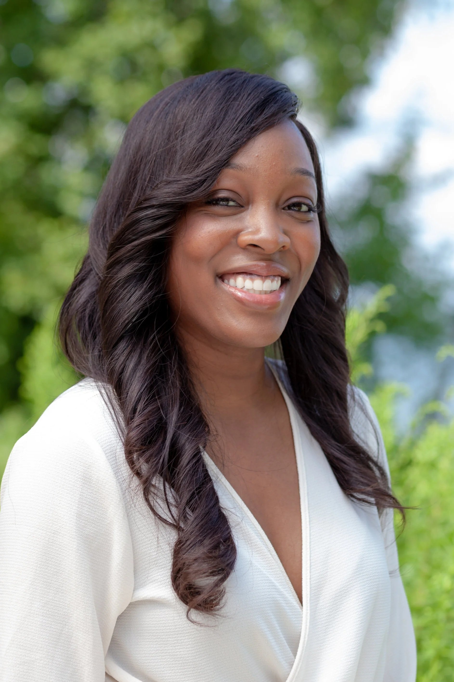 A woman with dark, wavy hair smiling outdoors with a background of green trees and foliage.