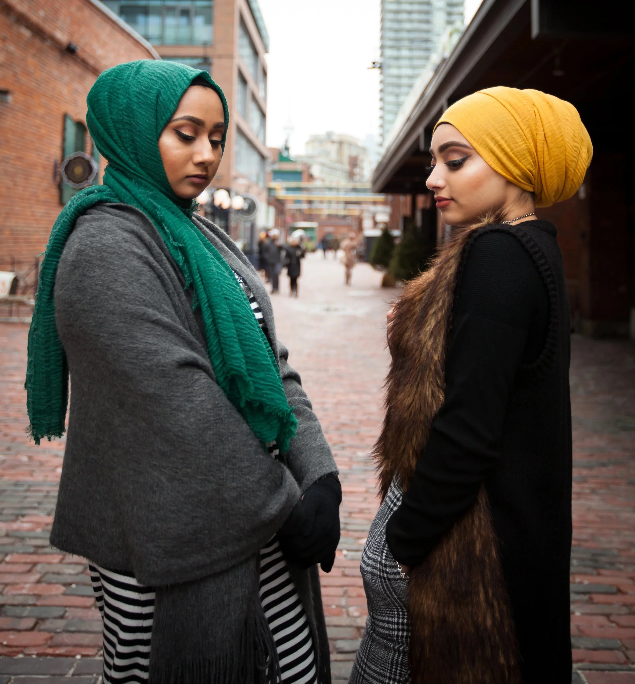 Two women with headscarves standing close together outdoors in an urban setting with brick and glass buildings, looking down with serious expressions.
