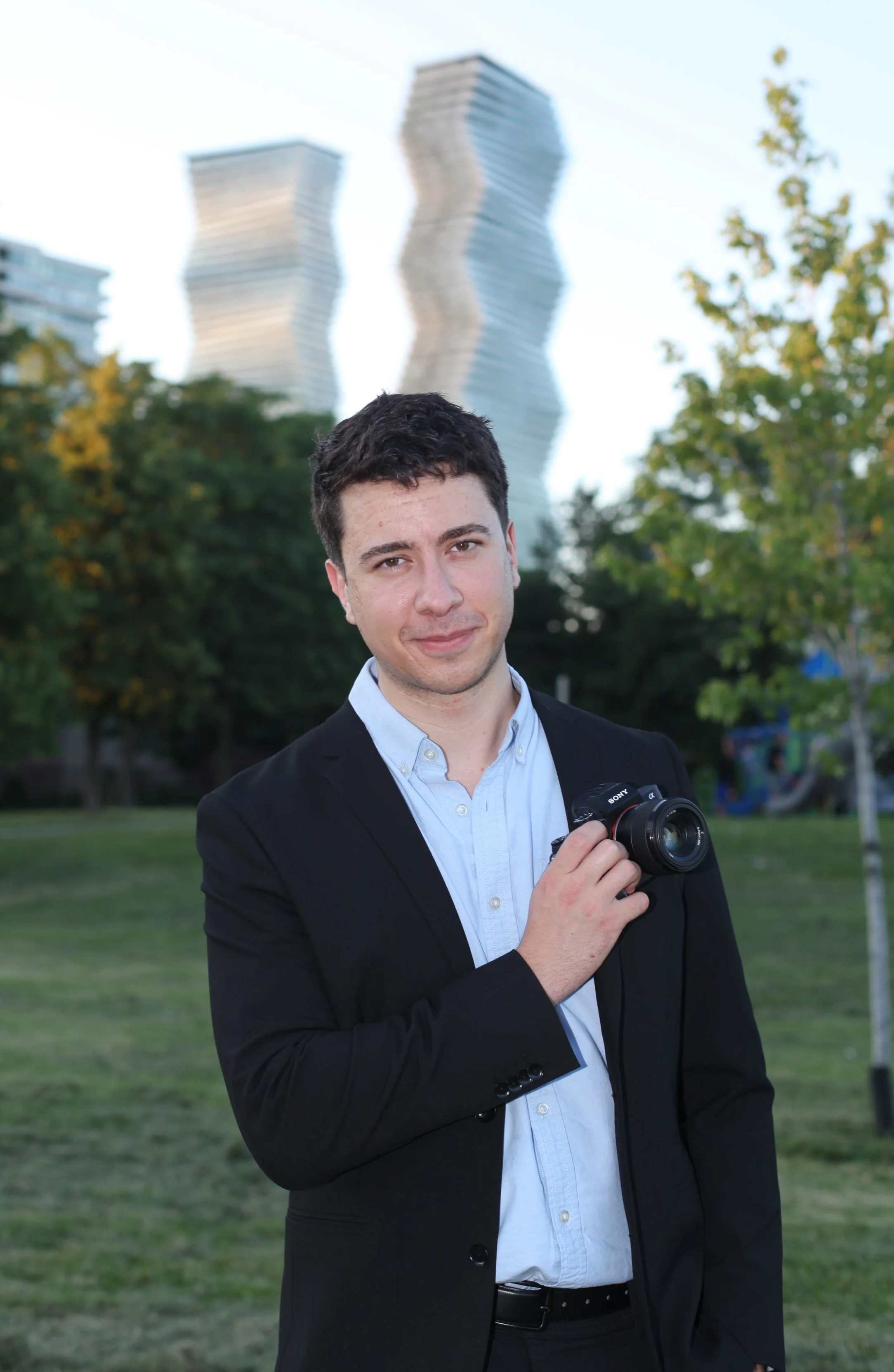 A young man in a dark suit holding a camera in a park with trees and a grassy area, with modern wavy skyscrapers in the background.