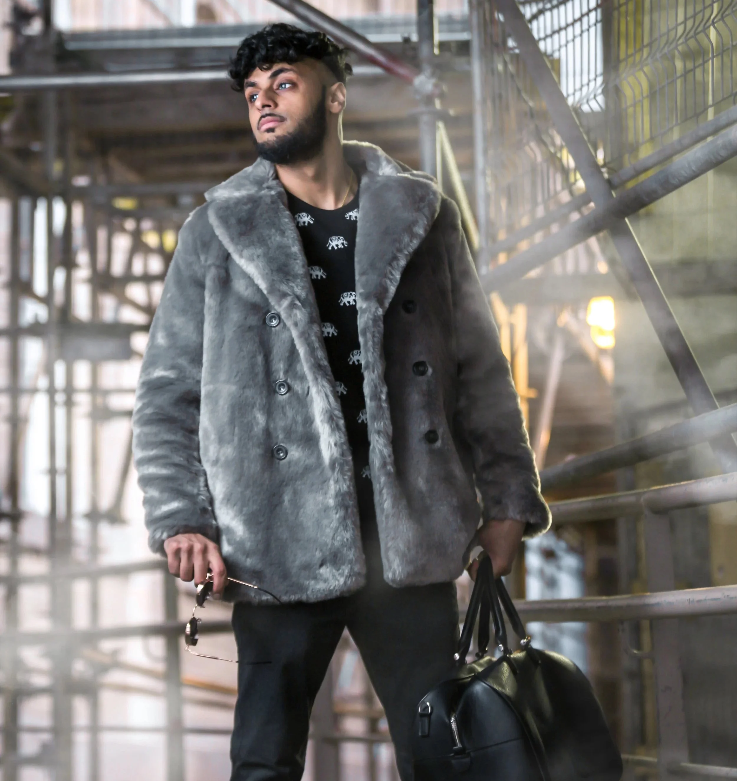 A young man with a beard and curly hair standing in an industrial or warehouse setting with metal scaffolding. He is dressed in a gray fur-lined coat over a black shirt with white elephant patterns and is holding a face mask and a black bag.