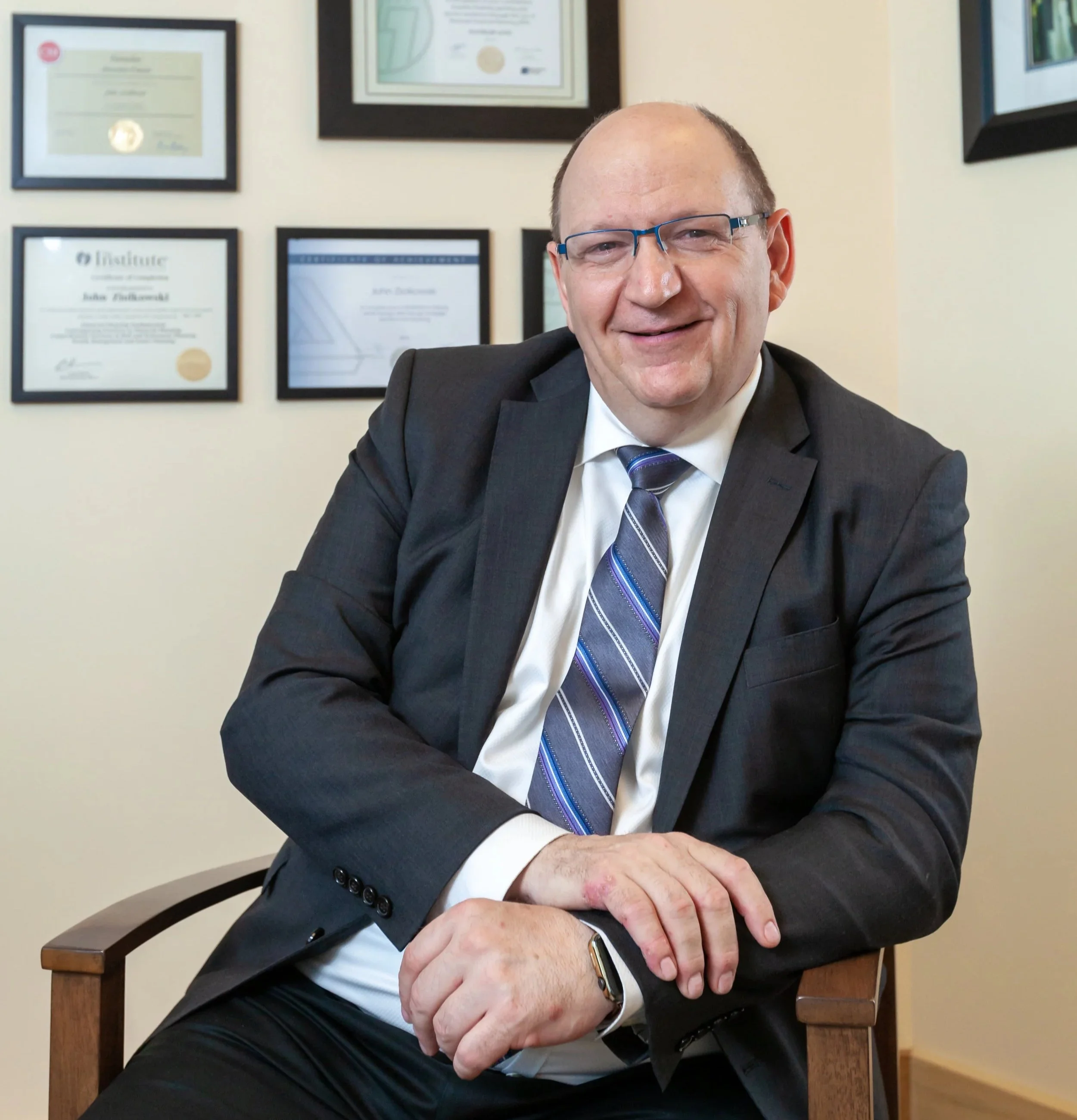 A man dressed in a suit and tie sitting in a wooden chair, smiling, with framed certificates on the wall behind him.