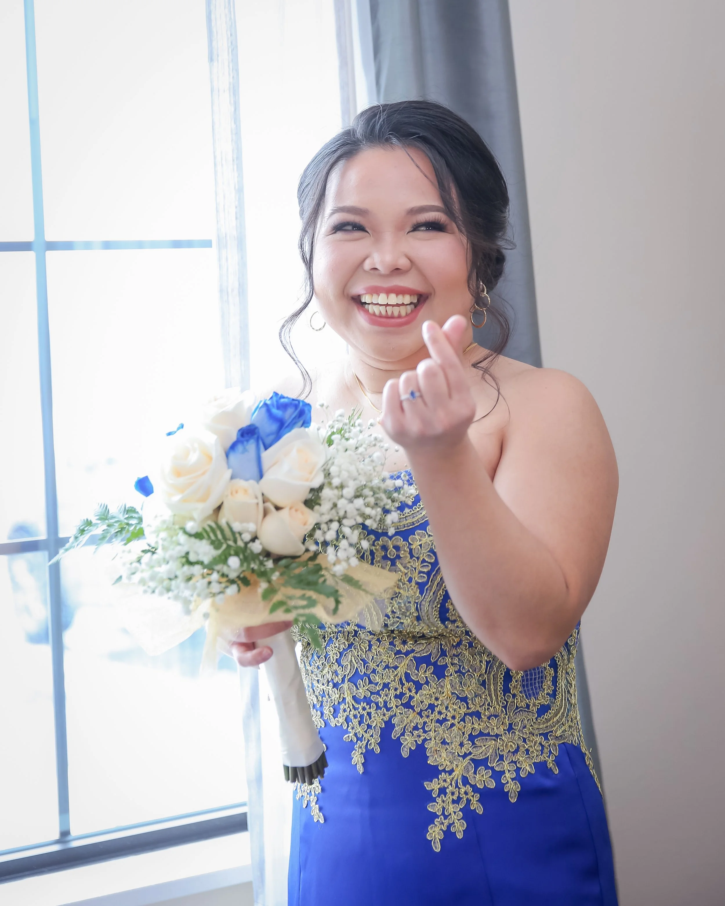 A woman smiling and making a heart gesture with her fingers while holding a bouquet of white and blue flowers. She is wearing a blue dress with gold embroidery and standing near a window with curtains.