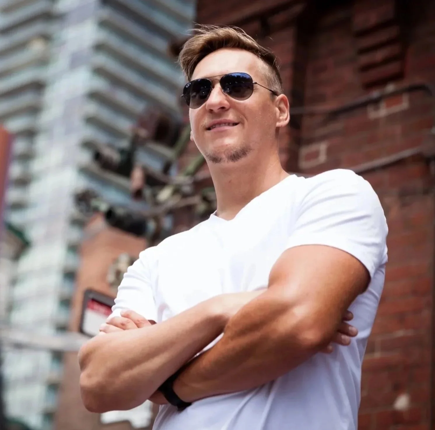A man with short brown hair, sunglasses, and a goatee is standing outdoors with his arms crossed, smiling slightly, in front of a brick building and some structures.