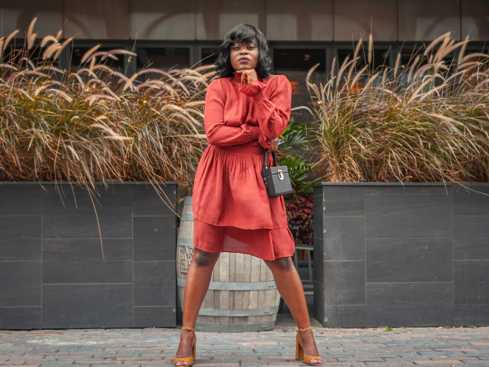 Woman standing outdoors in front of plants and a modern building, wearing a red dress, high-heeled shoes, holding a small black handbag, and striking a confident pose.