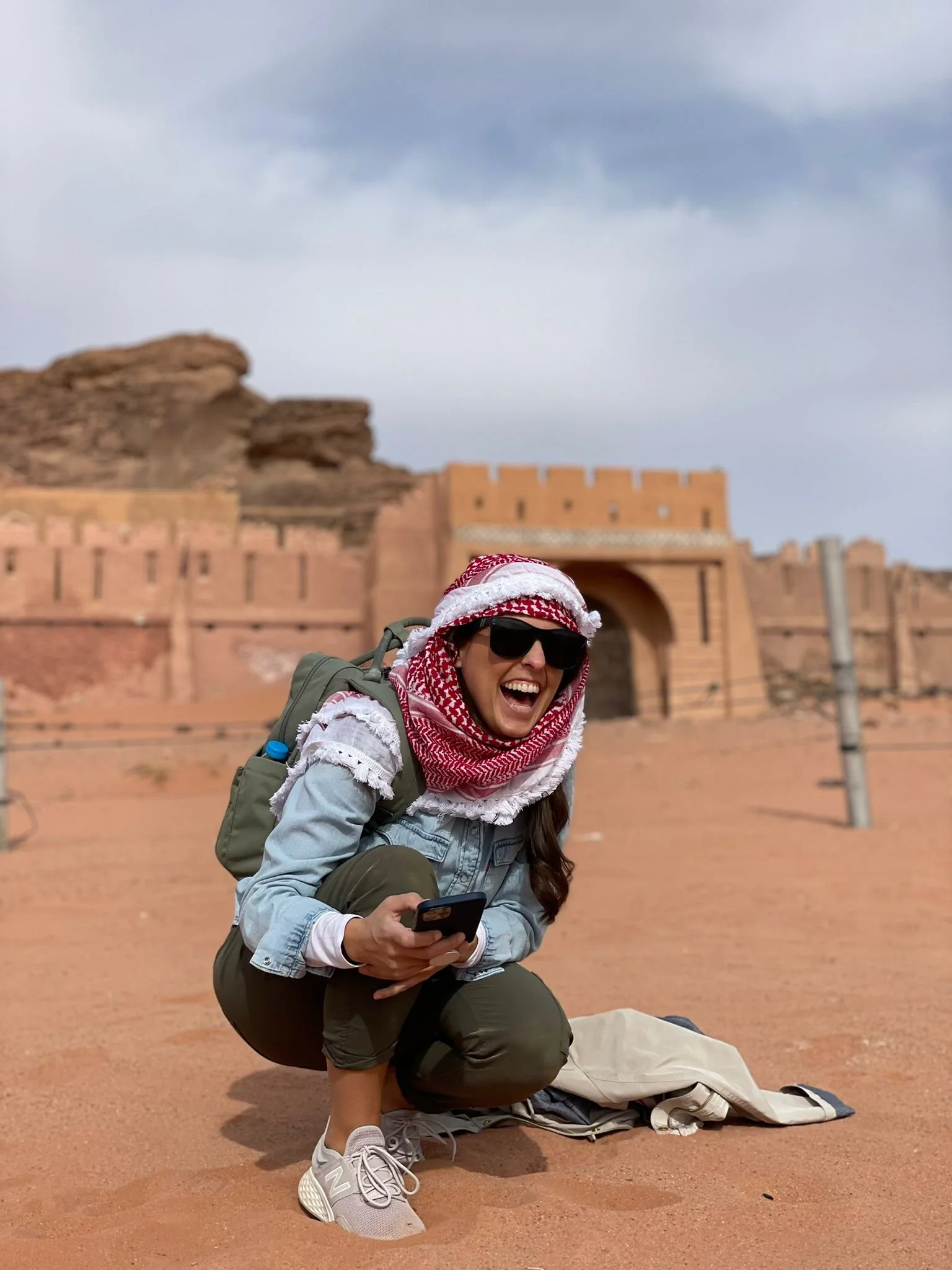 A woman crouching on sandy ground, smiling and holding a smartphone, with a desert city wall and archway in the background.