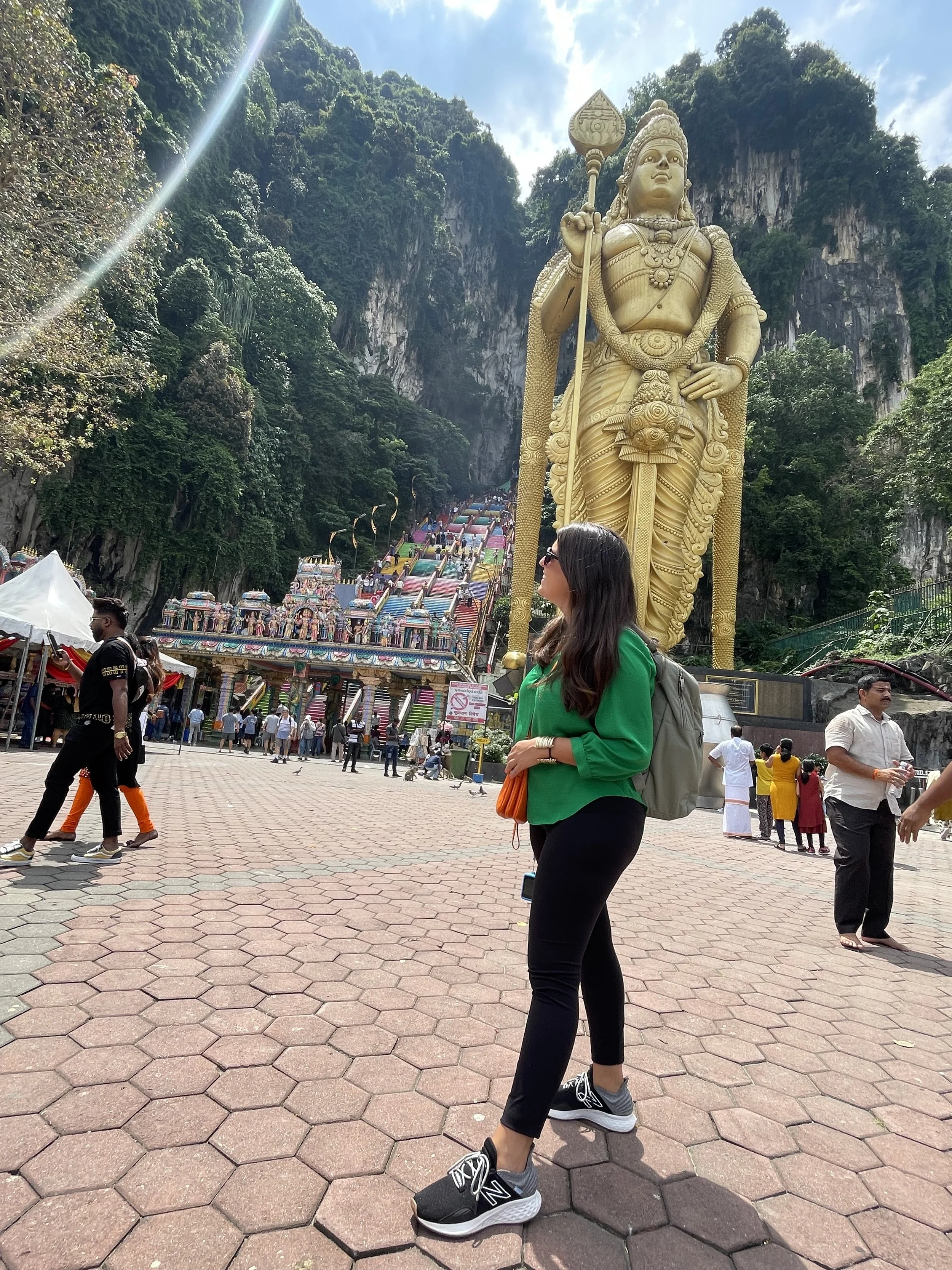 A woman standing outside Batu Caves in Malaysia, with a large golden statue of Lord Murugan and colorful steps leading up a mountainside in the background.