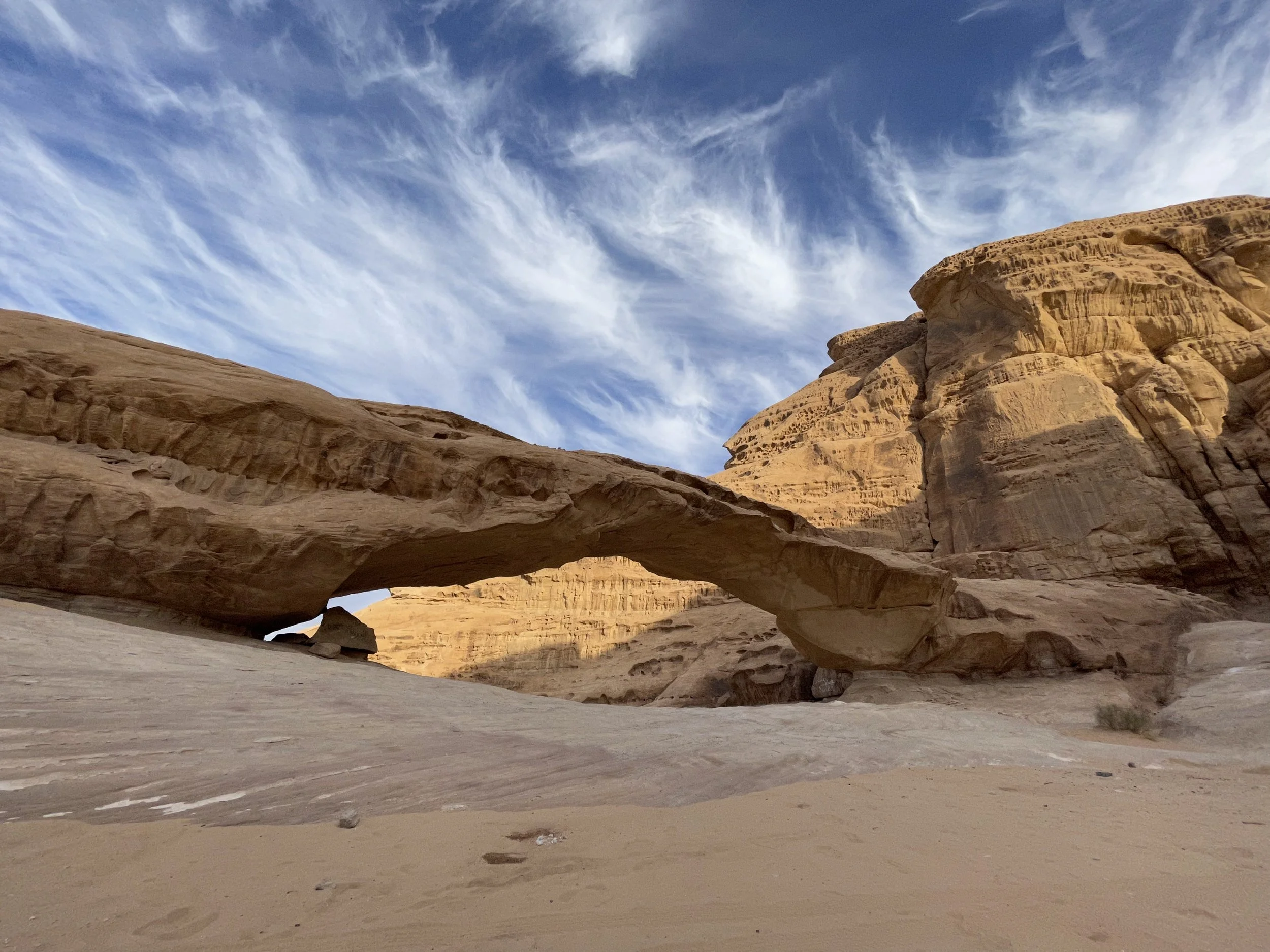 A natural rock formation in a desert landscape with a large arch-shaped rock and sandy ground, under a blue sky with wispy clouds.