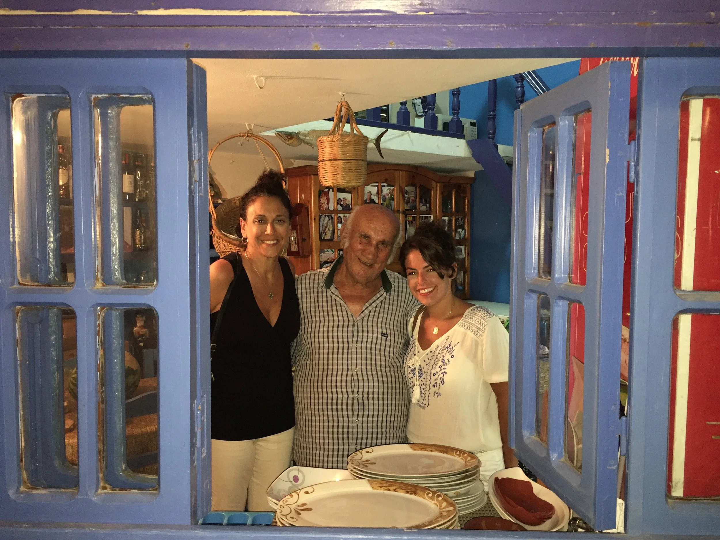 Three people standing together inside a restaurant, smiling and posing for the photo. The image is taken through a window with blue shutters.