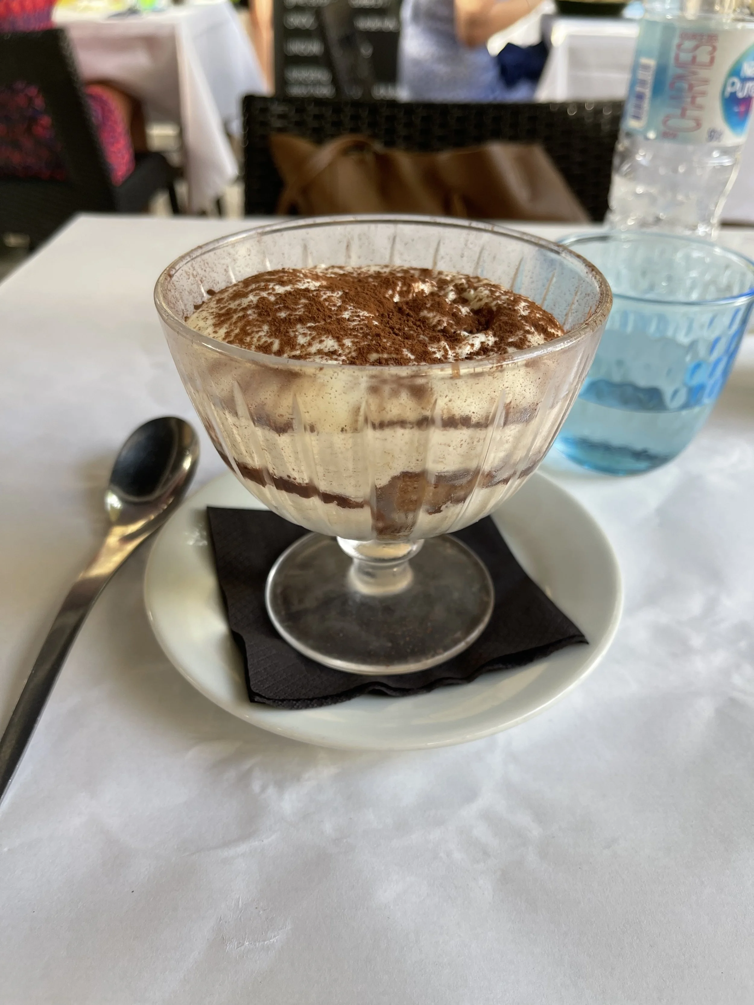 A glass bowl of tiramisu dessert topped with cocoa powder, placed on a black napkin on a white plate with a spoon, on a white table.