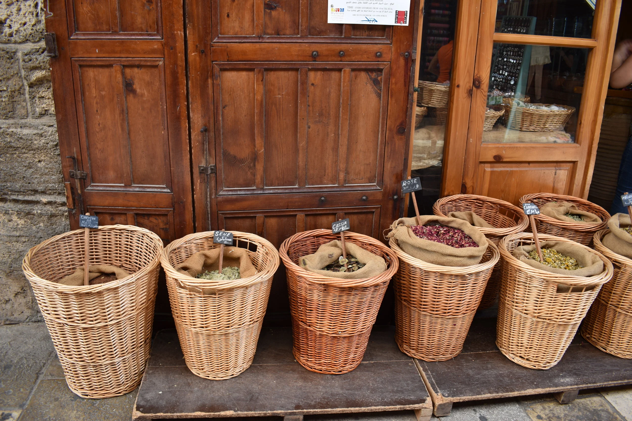 Six baskets of dried herbs and flowers placed on a wooden shelf outside a store with a wooden door.