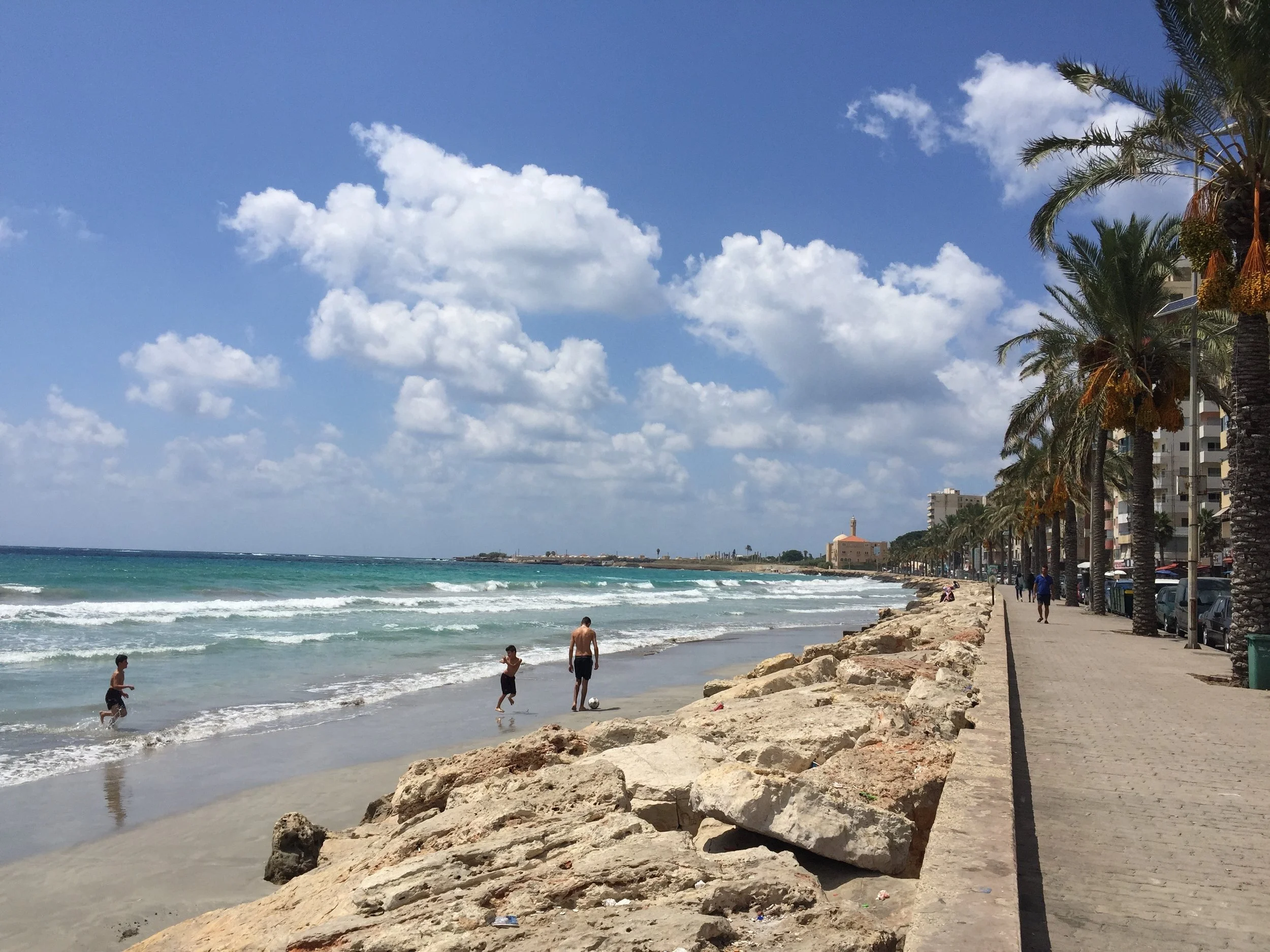 People playing near the shoreline of a beach with palm trees and high-rise buildings along the coast under a partly cloudy sky.