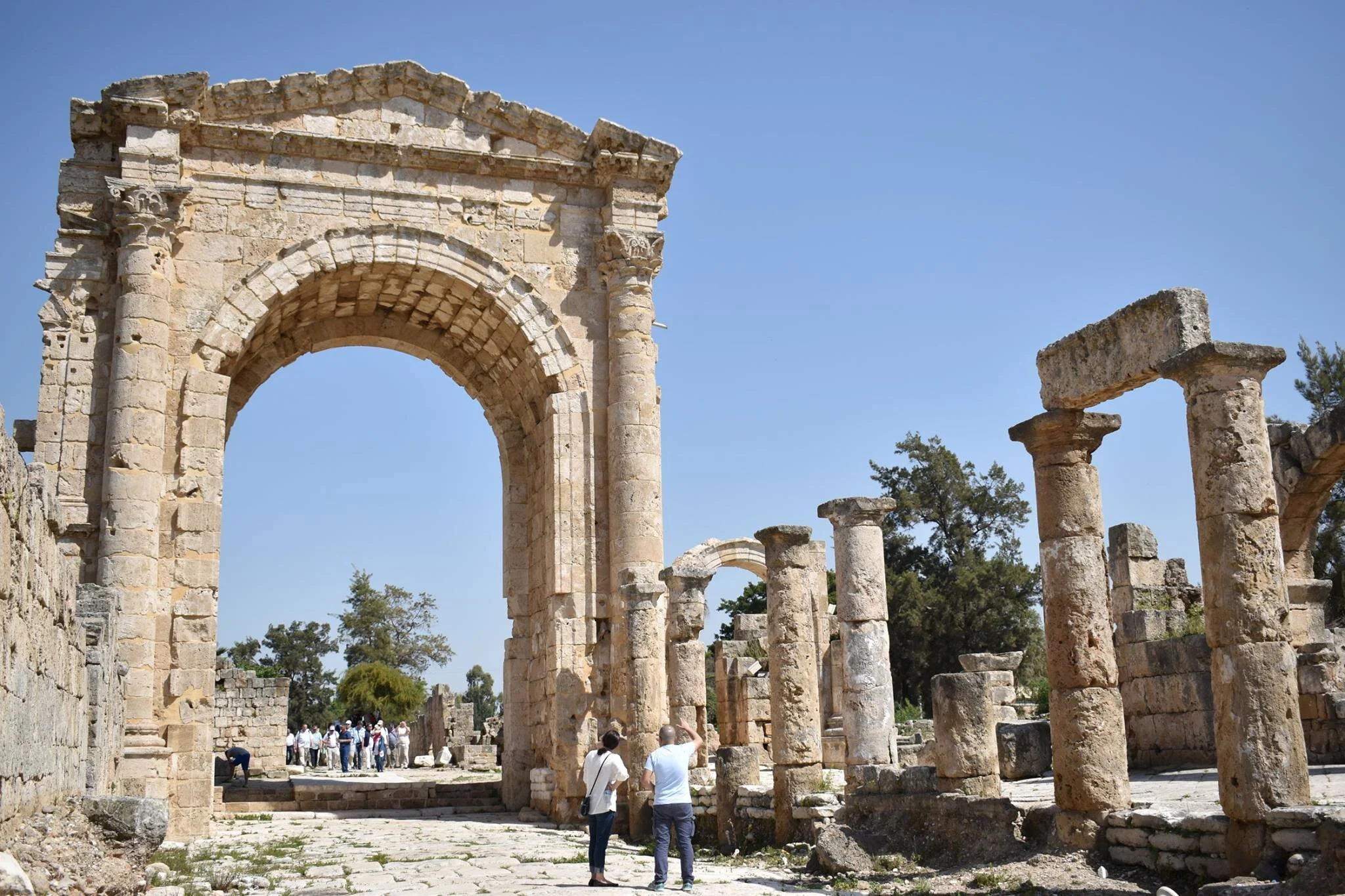 Ancient stone ruins with tall columns and an archway, some visitors exploring the site on a sunny day.