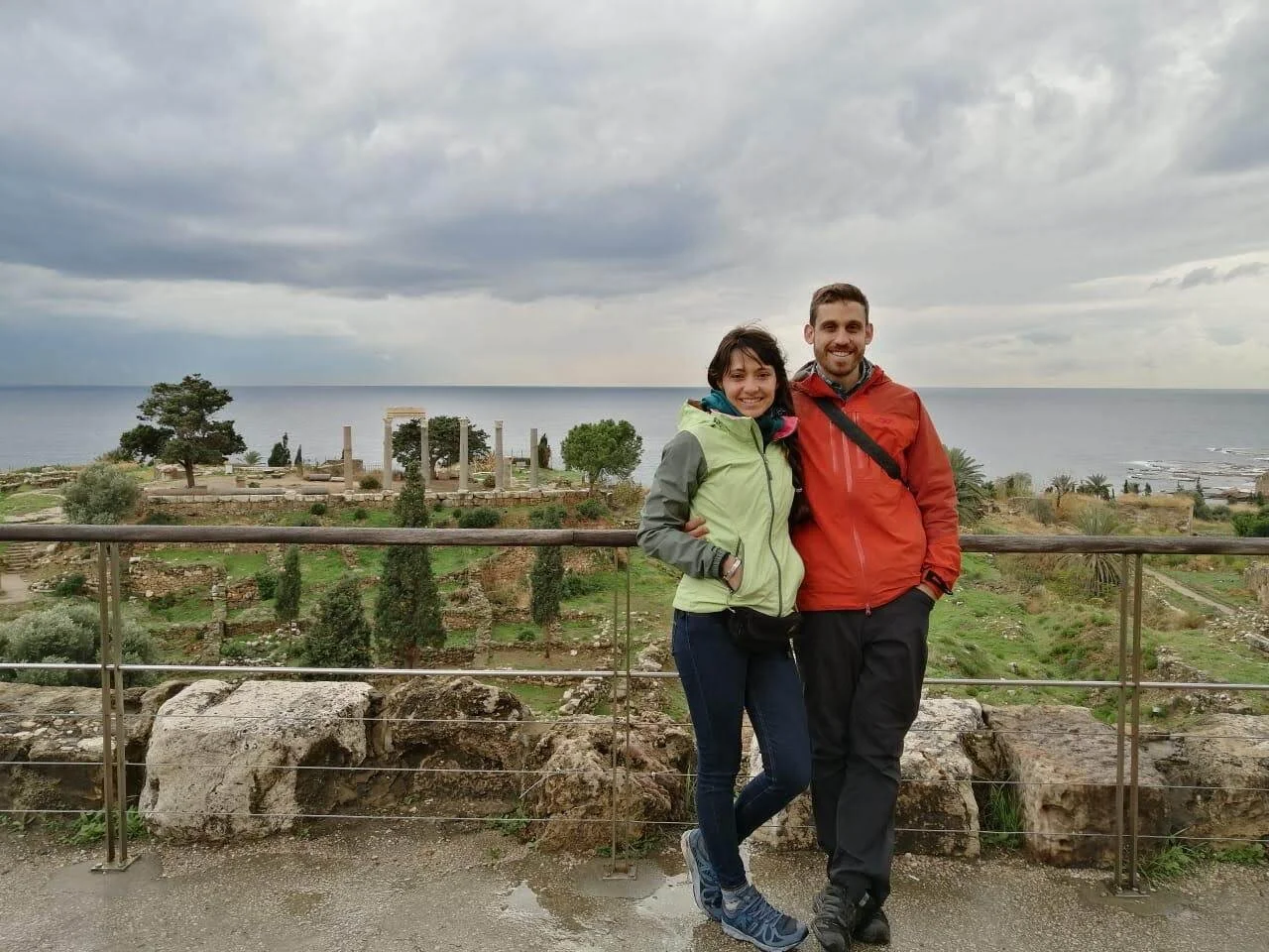 A smiling couple standing arm-in-arm outdoors with a historic site and the ocean in the background on a cloudy day.