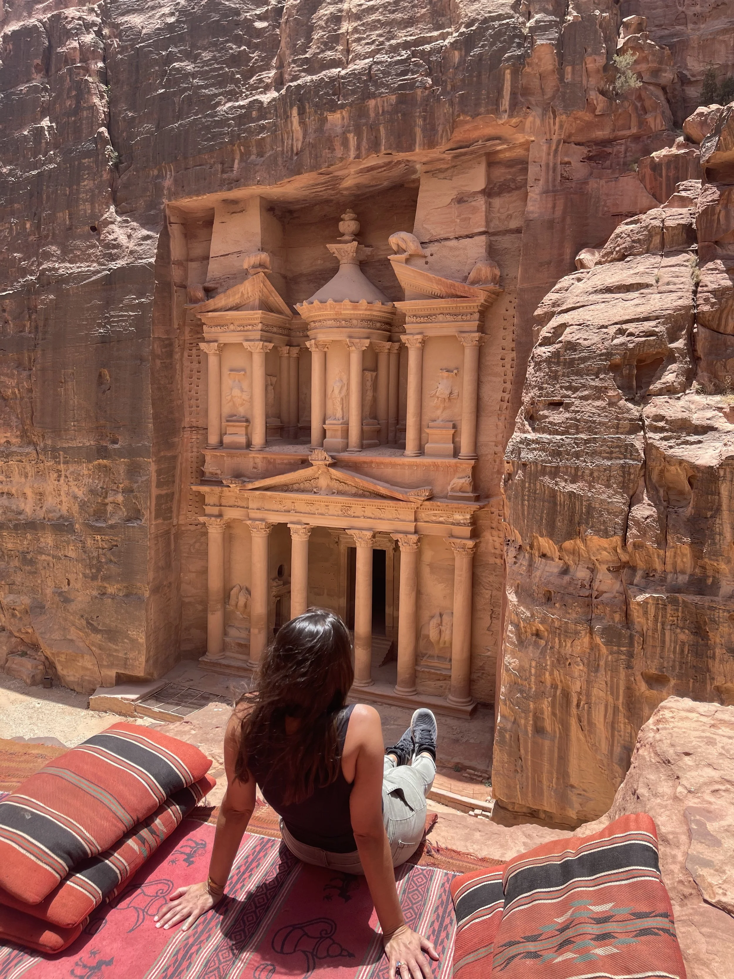 A woman sitting on a cushion overlooks the ancient rock-carved city of Petra in Jordan, specifically the Treasury building carved into the sandstone cliffs.