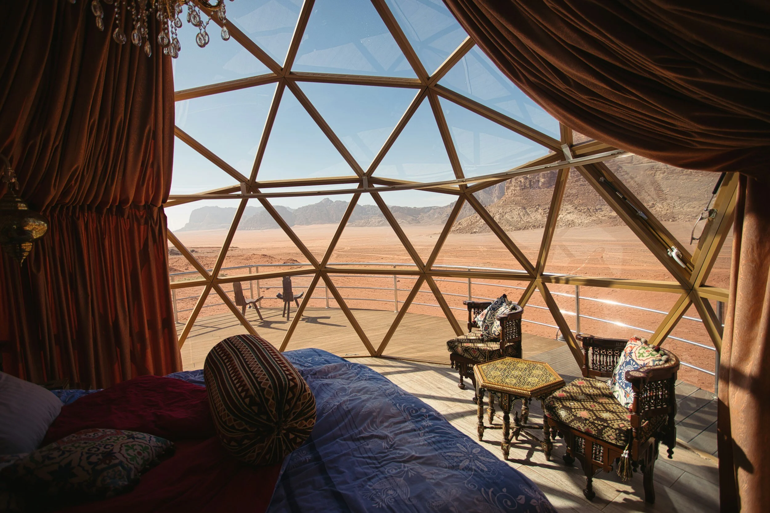 Interior view of a geodesic dome structure with a bed, ornate pillows, and curtains, overlooking a desert landscape with mountains in the distance.