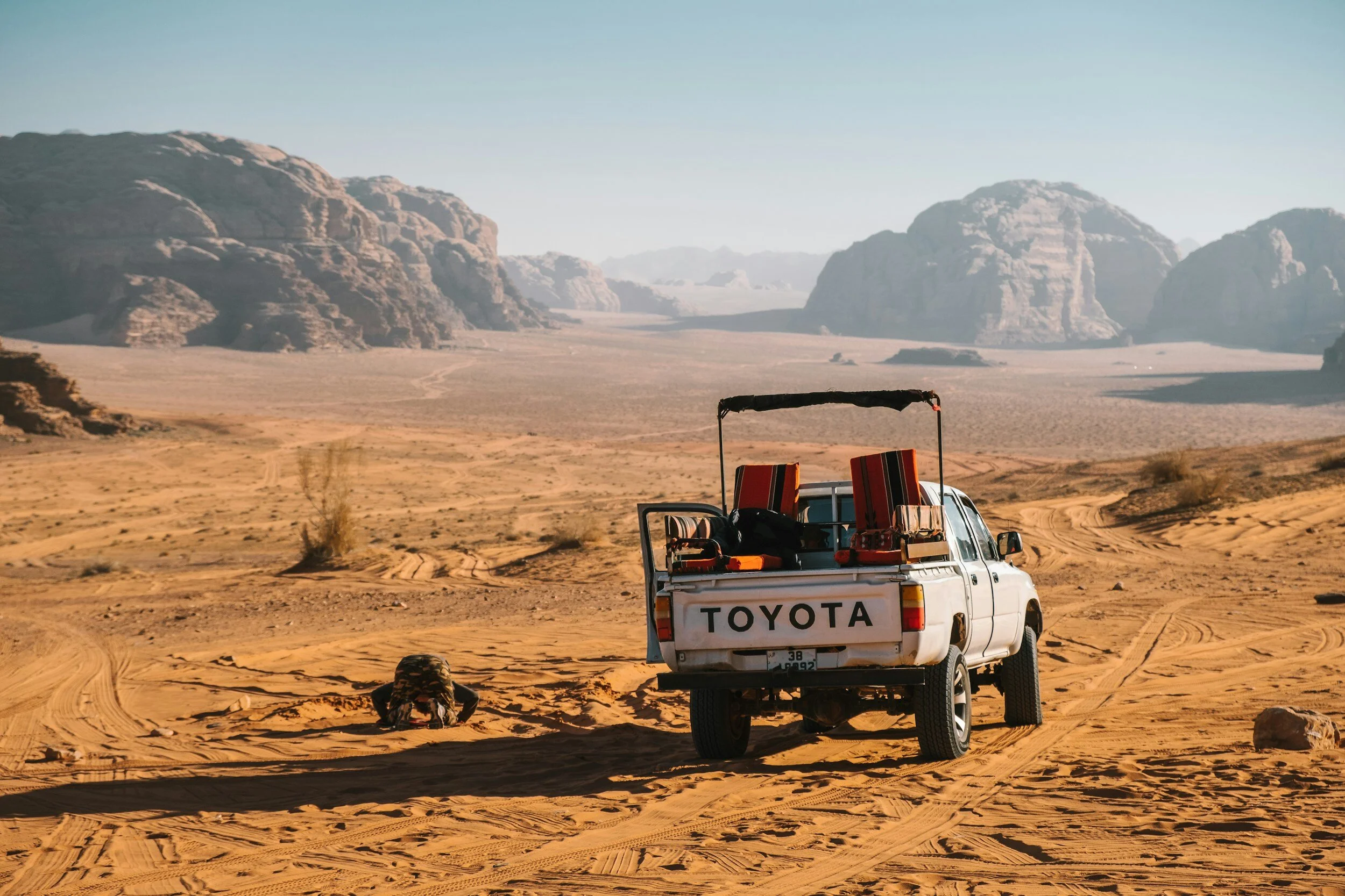A white Toyota pickup truck parked on a sandy desert landscape with a person crawling on the ground near it. The truck's bed contains two striped lounge chairs and a few bags, with rocky mountains in the background under a clear sky.
