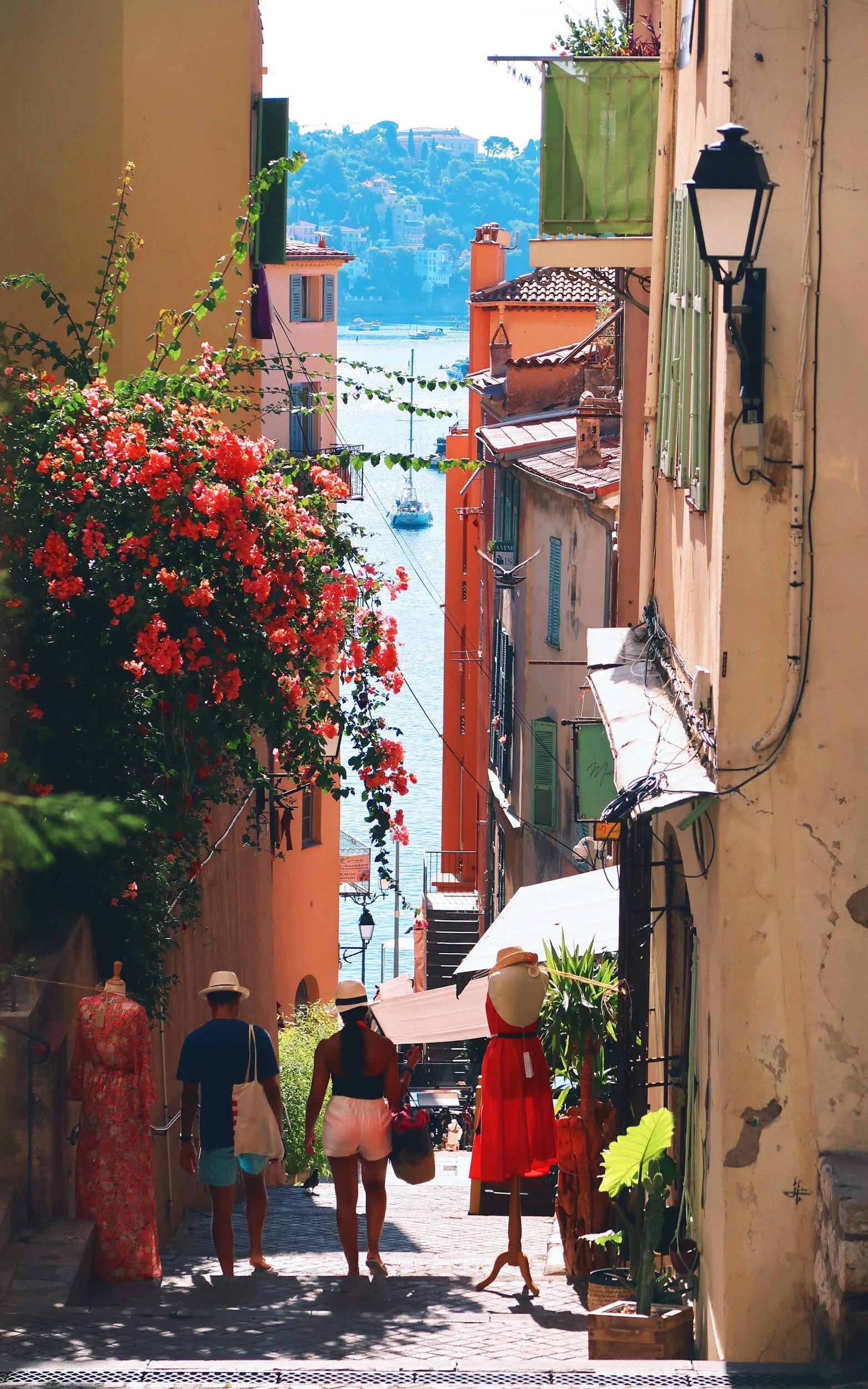 A vibrant alleyway leading to a waterfront with boats, flanked by colorful buildings, adorned with flowers, with three people walking away from the camera.