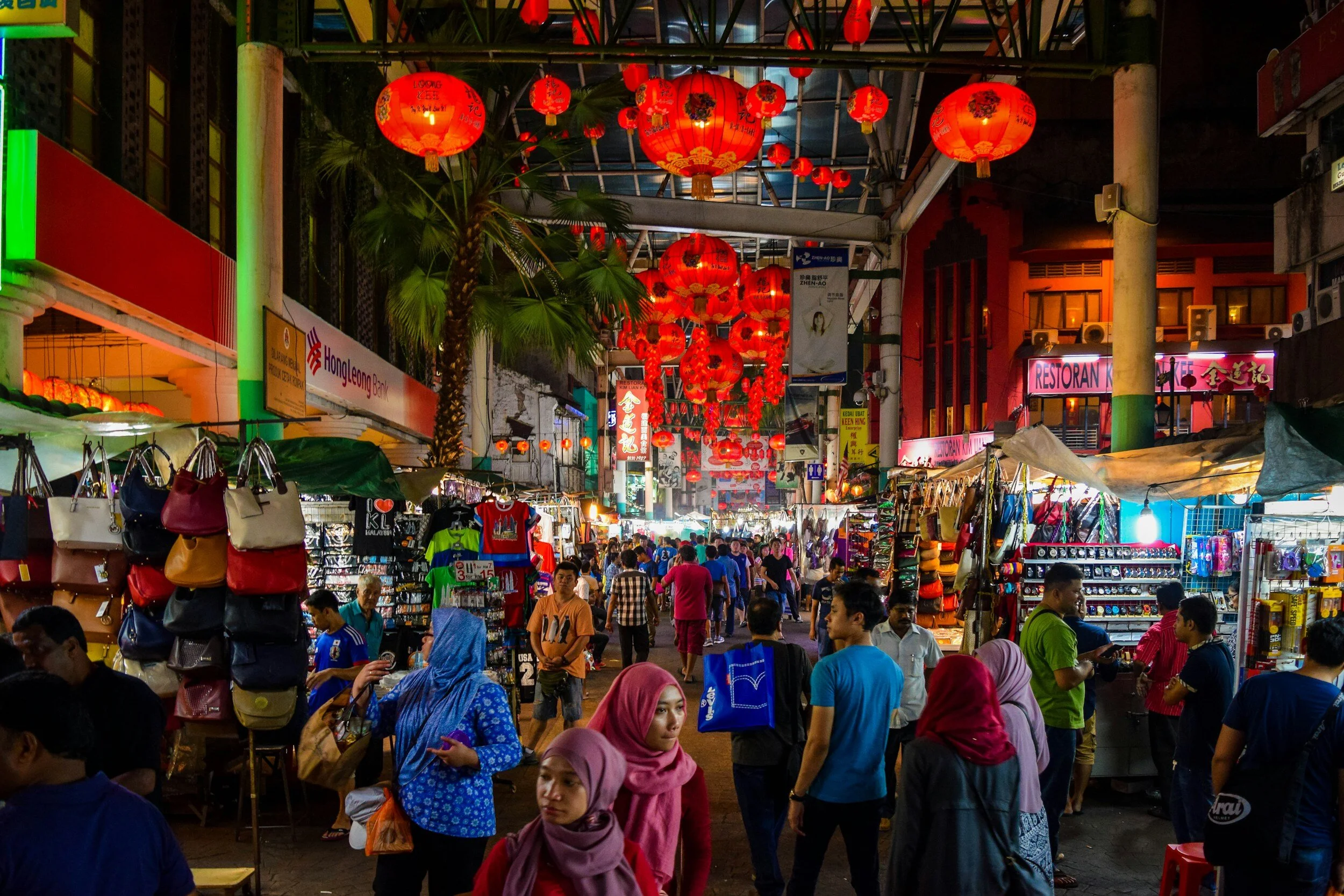 Night street market decorated with hanging red lanterns, with stalls selling bags, accessories, and clothing, and people walking and shopping.