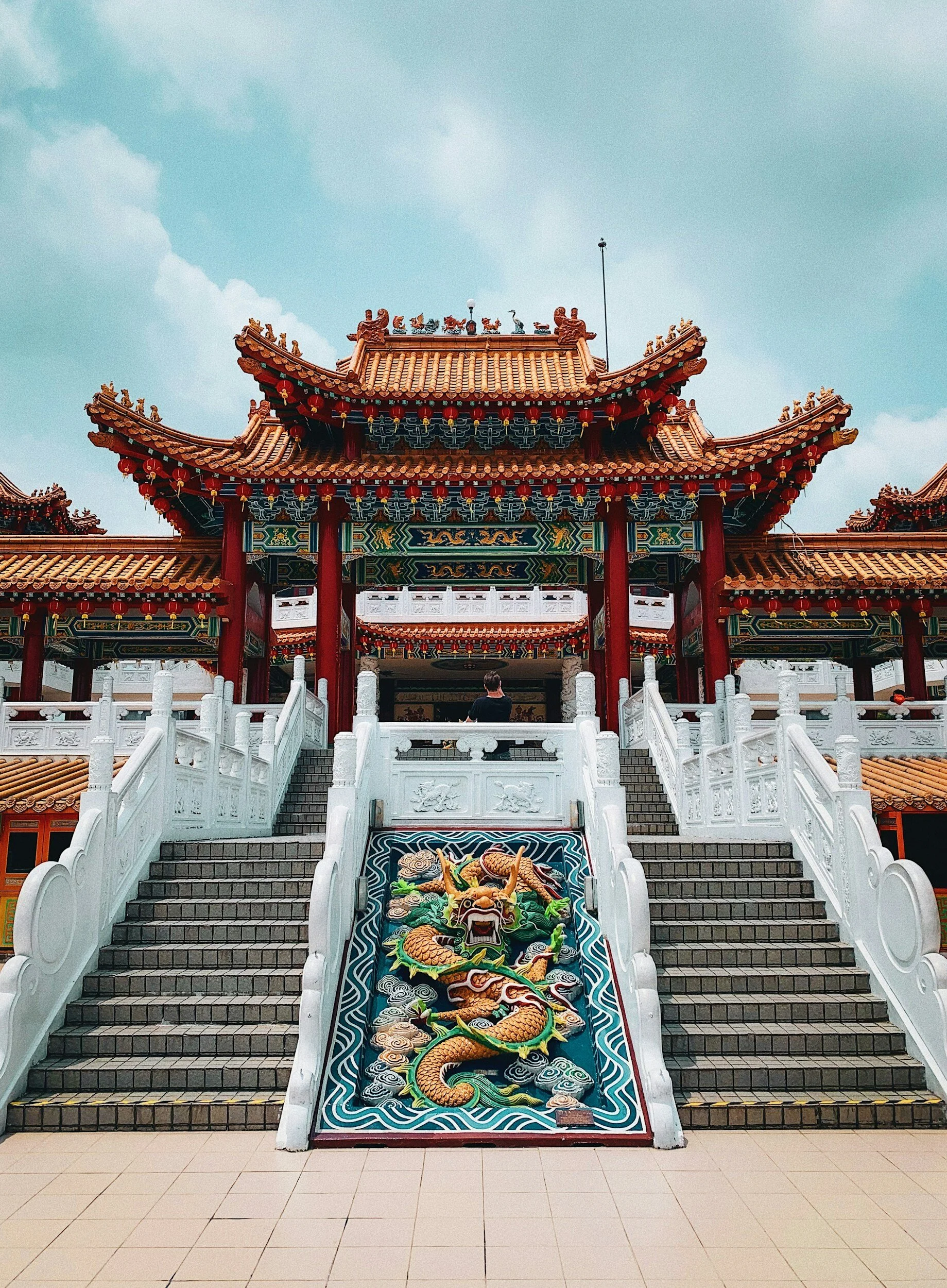 A traditional Asian temple with red, green, and gold ornate decorations, steep steps leading up to the entrance, and a colorful dragon sculpture on the stairs.
