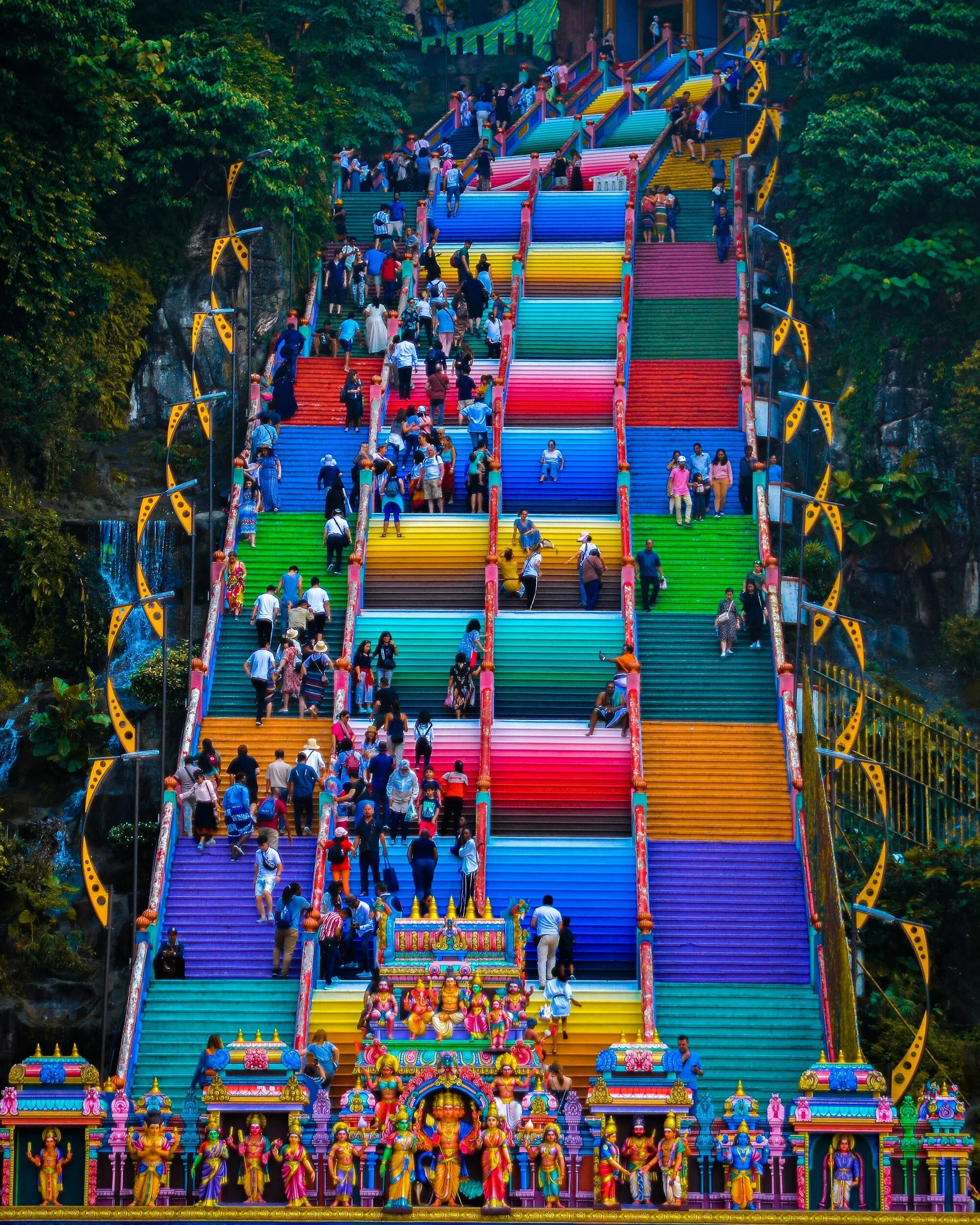 Colorful Hindu temple stairs with people walking up and down, surrounded by greenery and decorated with statues of deities at the bottom.
