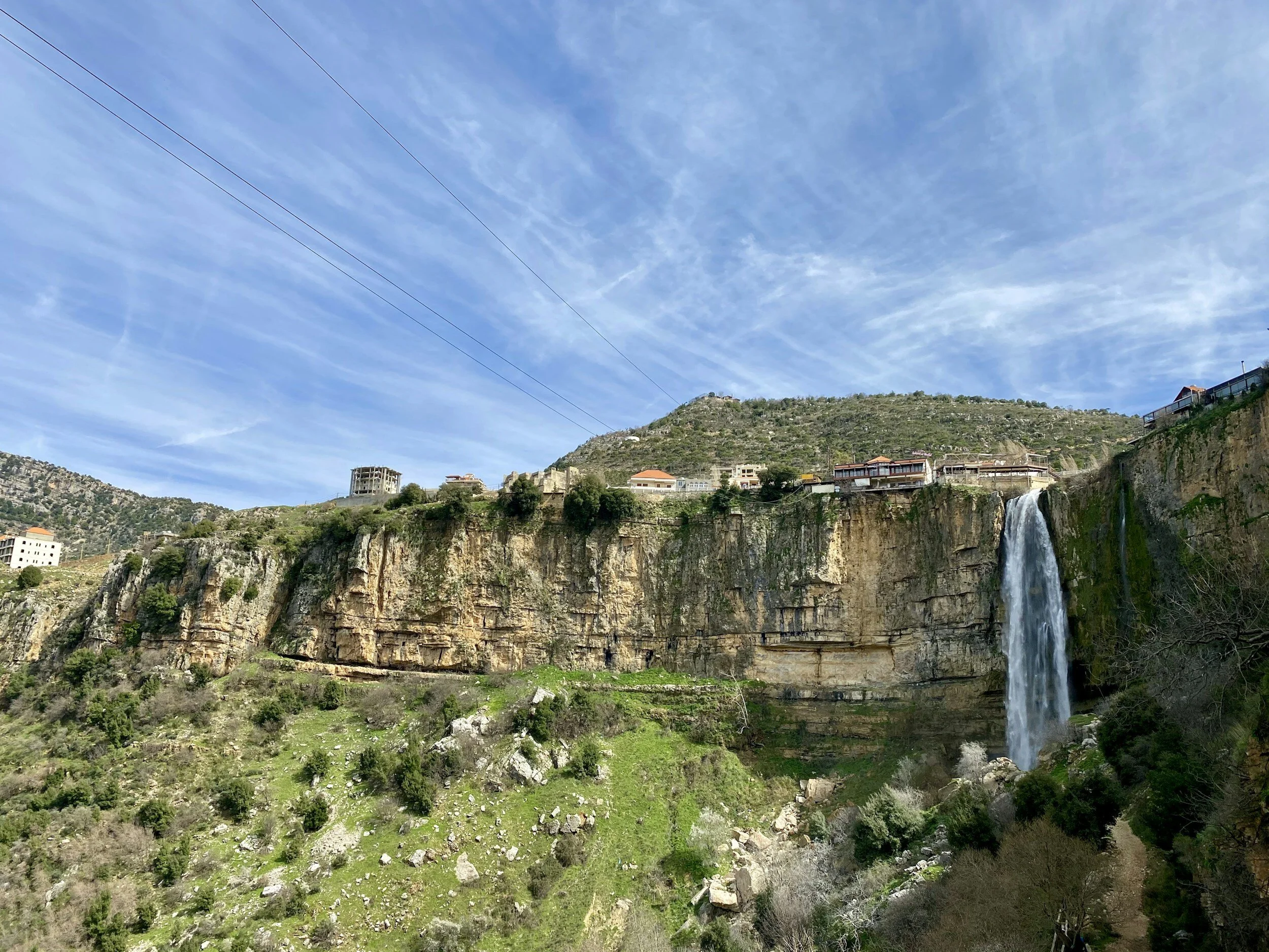 Scenic view of a waterfall flowing from a cliff into a lush green valley with houses on the hillside under a partly cloudy sky.