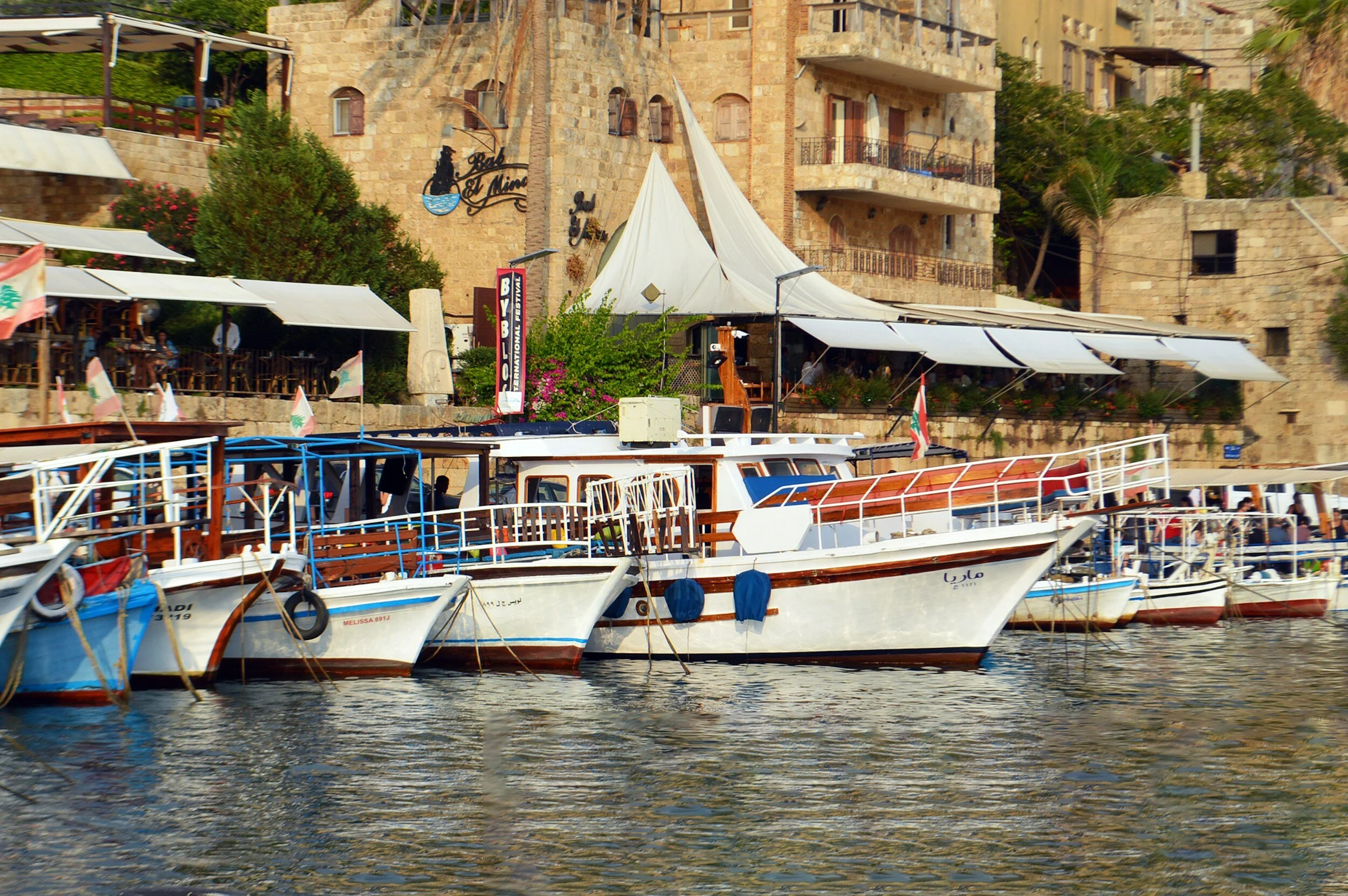 Boats docked at a waterfront restaurant with stone buildings and outdoor seating in the background.
