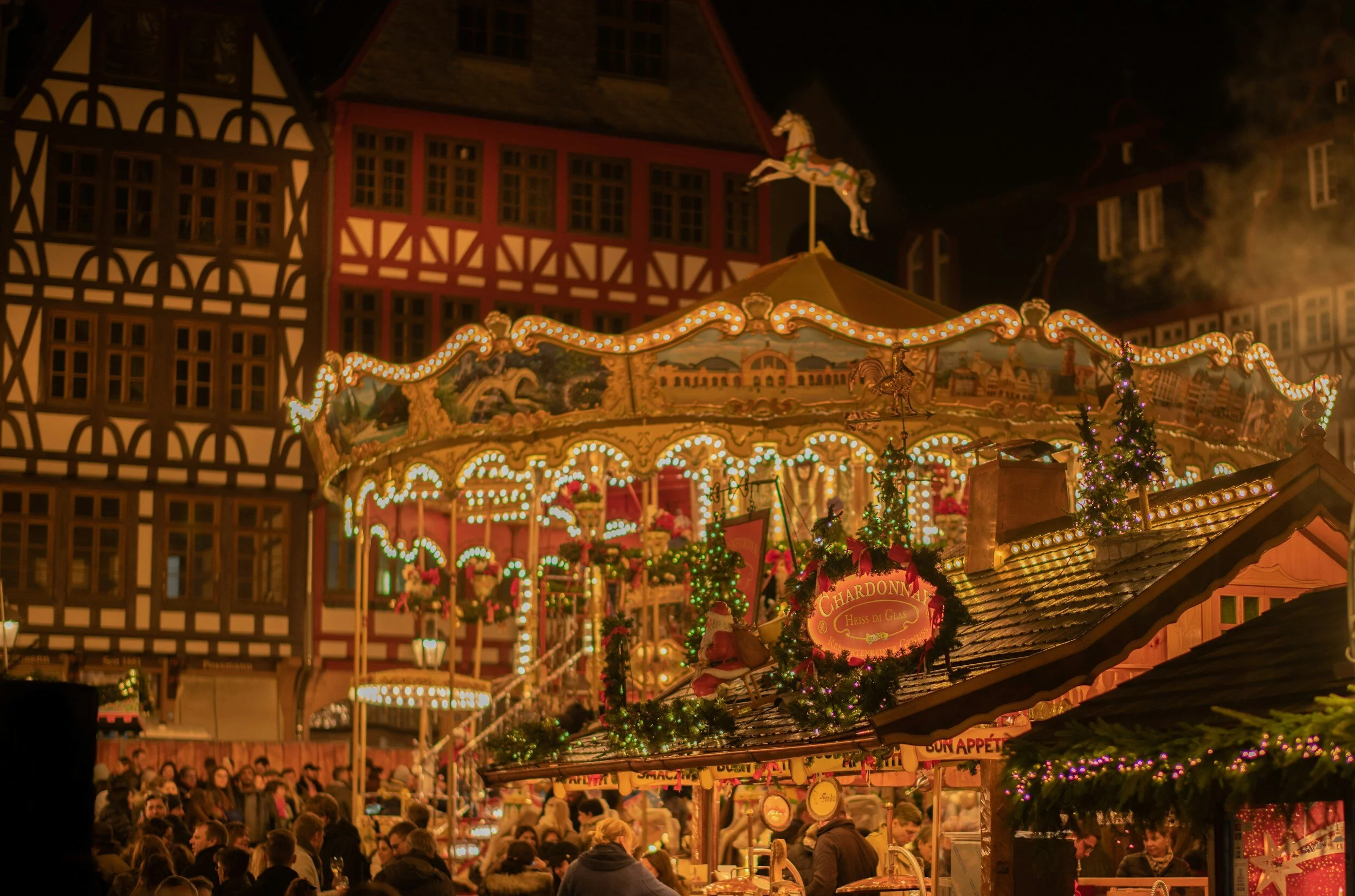 Christmas market at night with a carousel ride illuminated by lights, decorated with Christmas trees and festive decorations, surrounded by people.