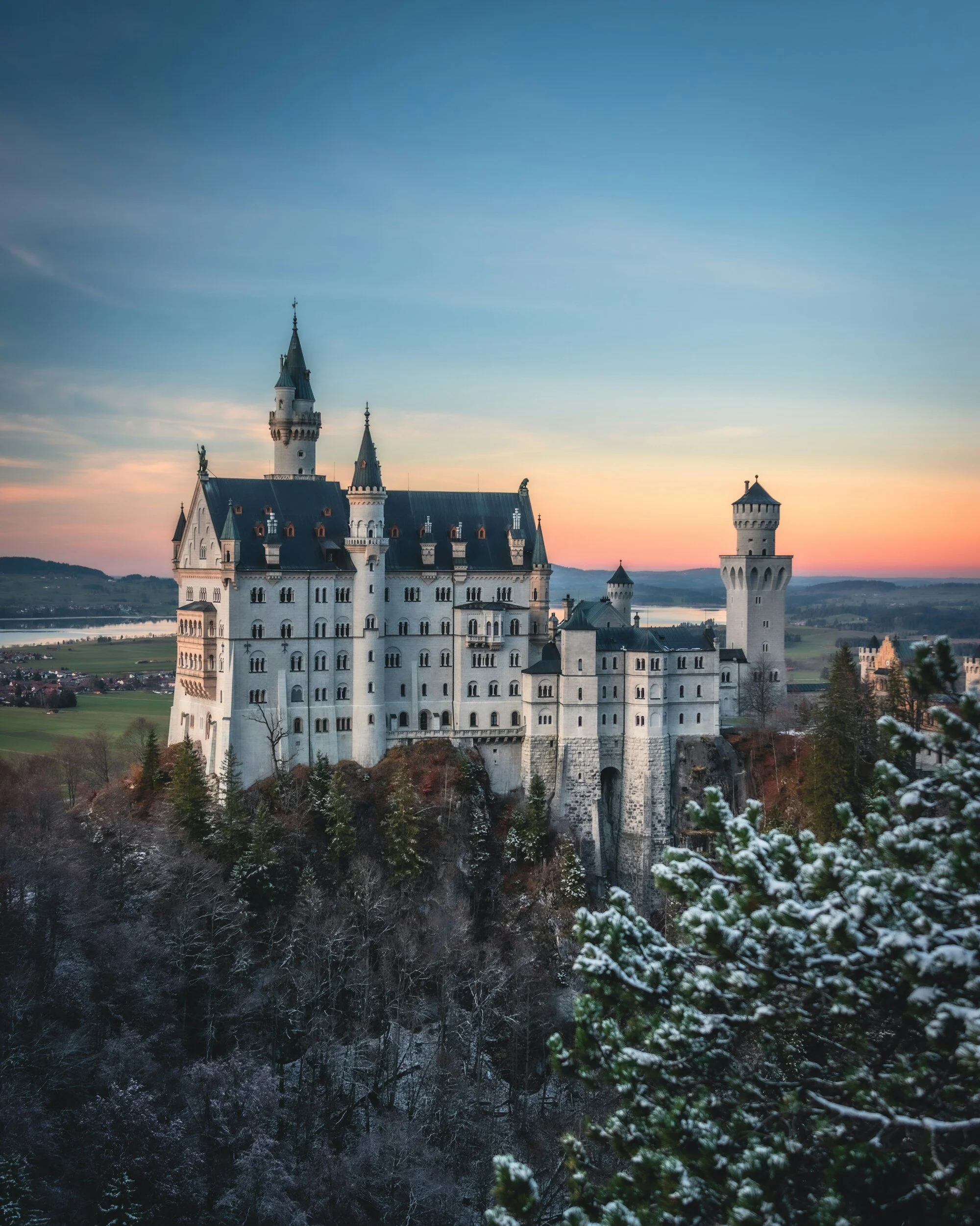 A large fairy tale castle with multiple towers and turrets, situated on a rocky hilltop beside a river, at sunset with a colorful sky.