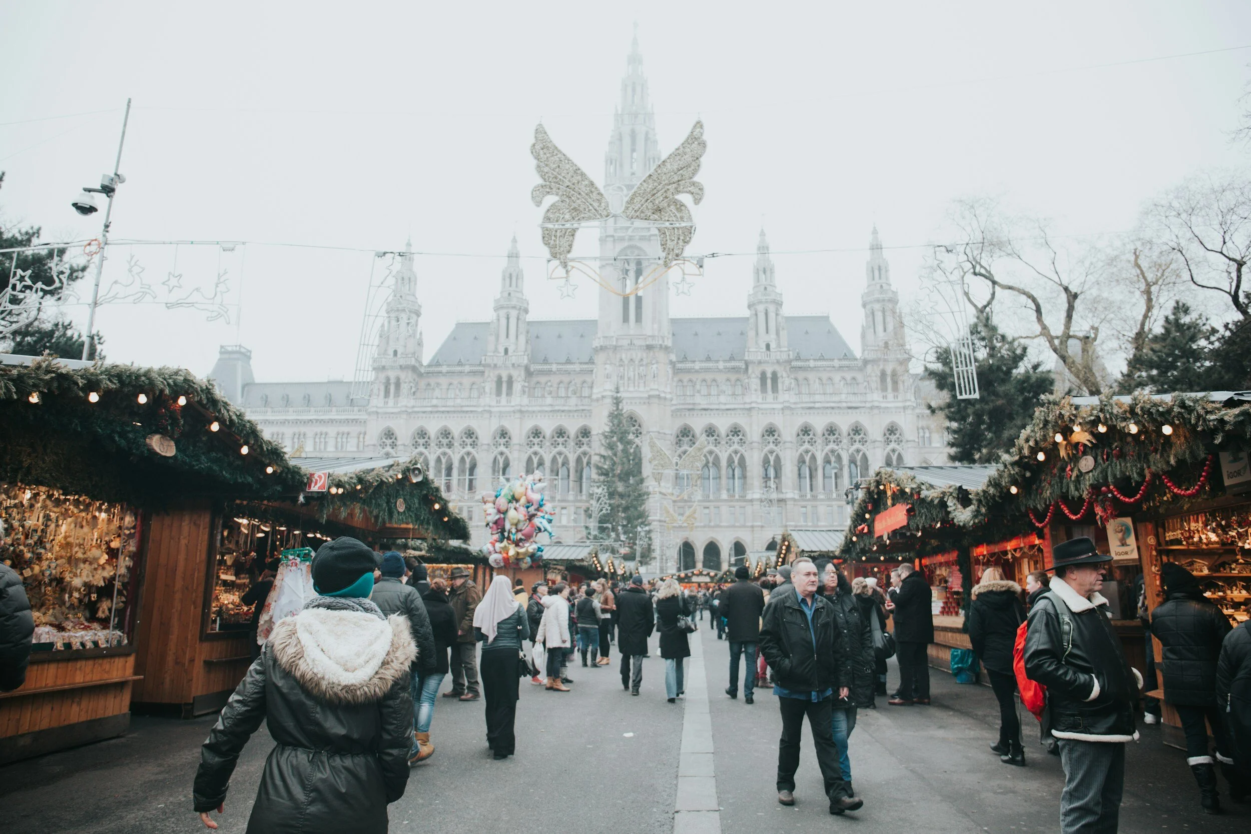 People shopping at a Christmas market in front of a historic gothic-style building with festive decorations, including a butterfly-shaped light fixture and balloon arrangements.
