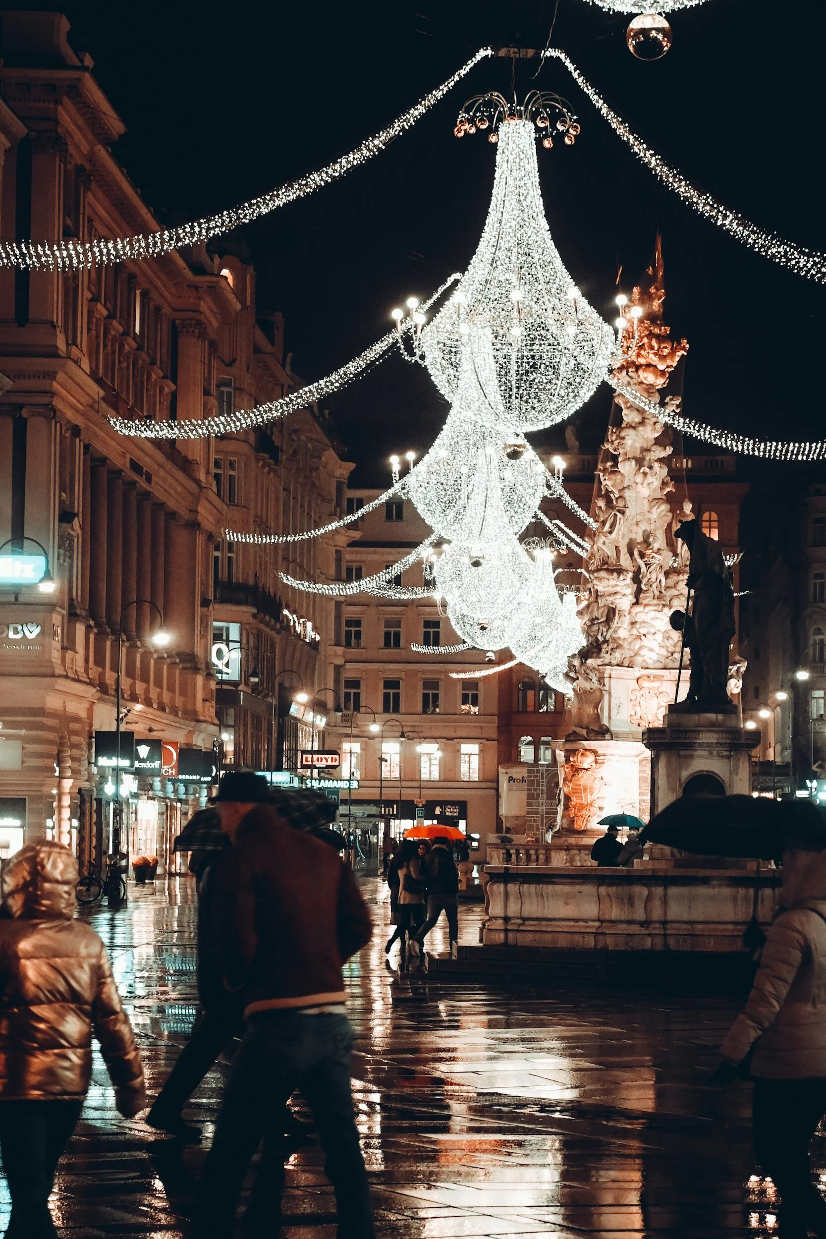 Nighttime city street with illuminated Weihnachtsmärkte (Christmas market) lights and decorations. People holding umbrellas walk on wet pavement near a stone statue and a large, ornate fountain, with historic buildings in the background.