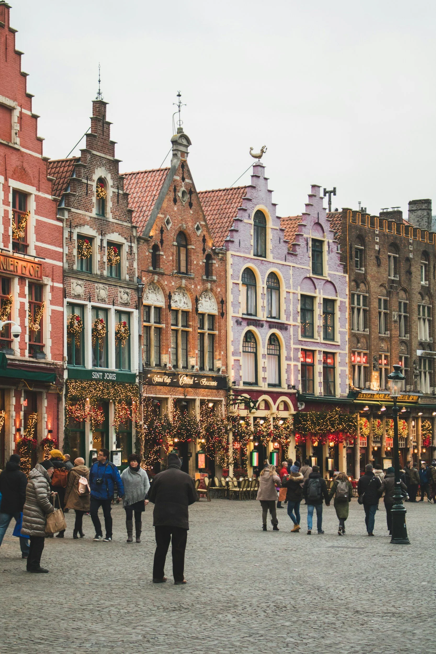European-style historic buildings decorated with Christmas lights and wreaths, with people walking and gathering in a cobblestone square during daytime.