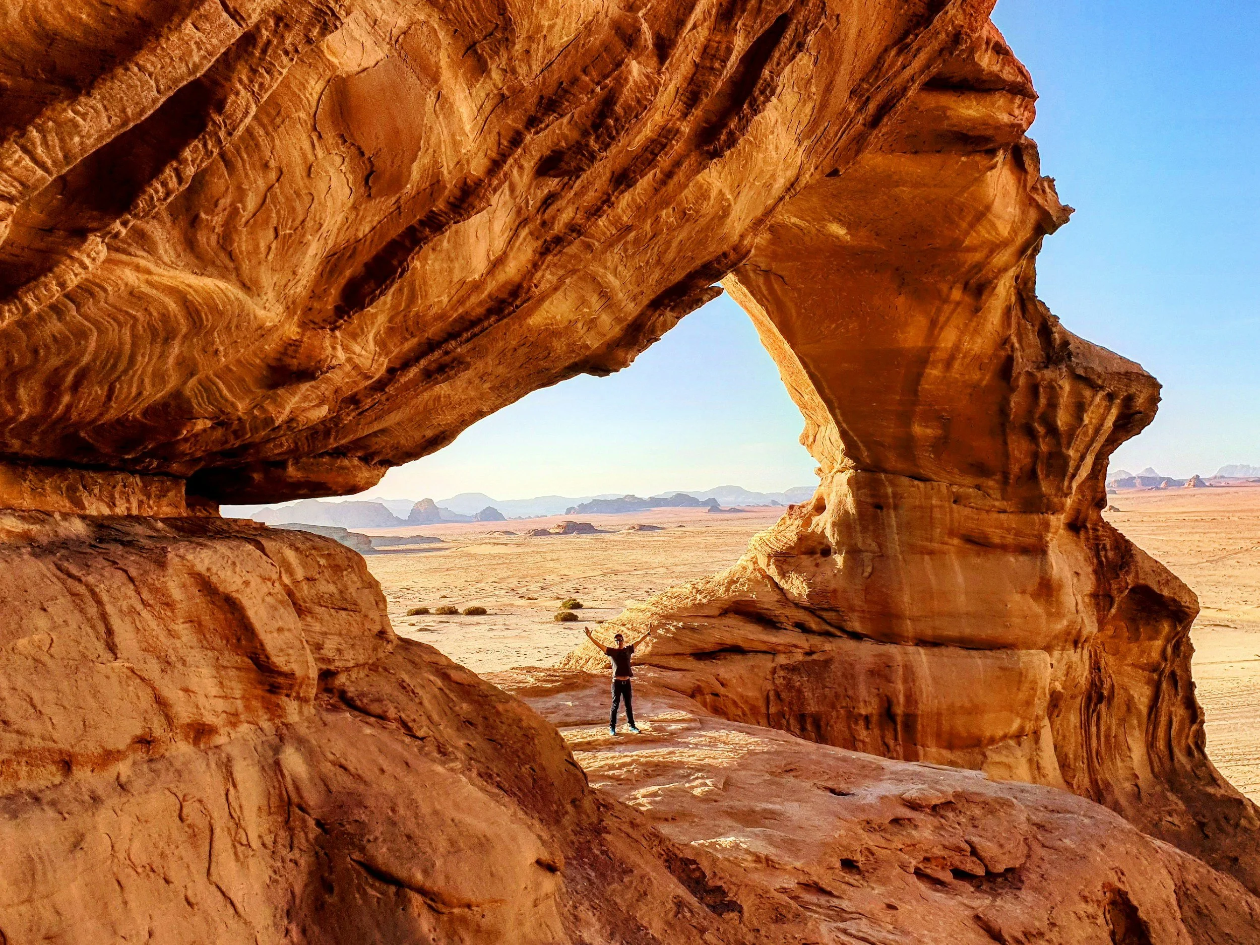 A person standing under a large natural rock arch formation in a desert landscape.