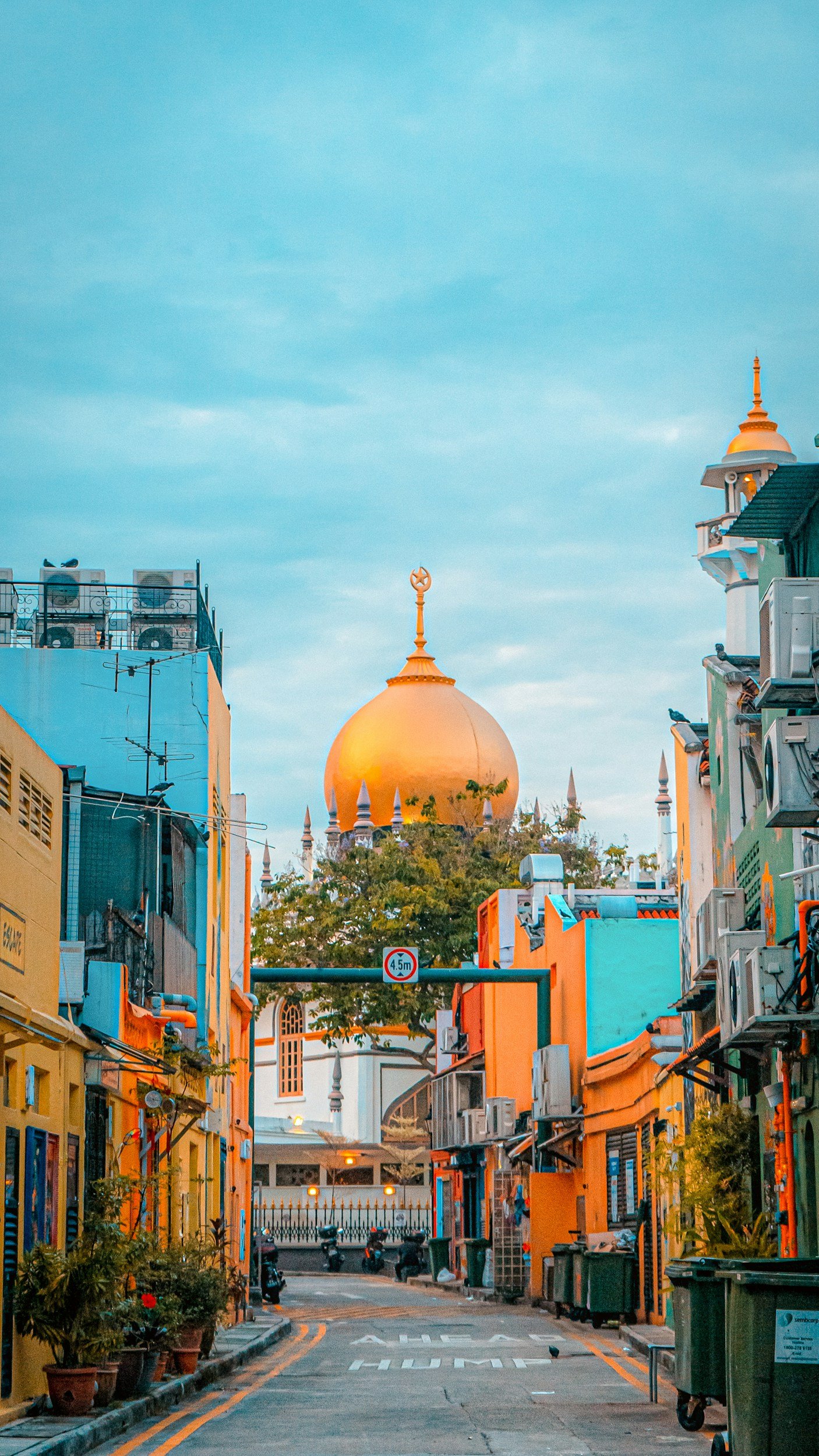 A street scene in an Asian city with colorful buildings, air conditioning units, and a gold-domed temple with spires in the background under a cloudy sky.
