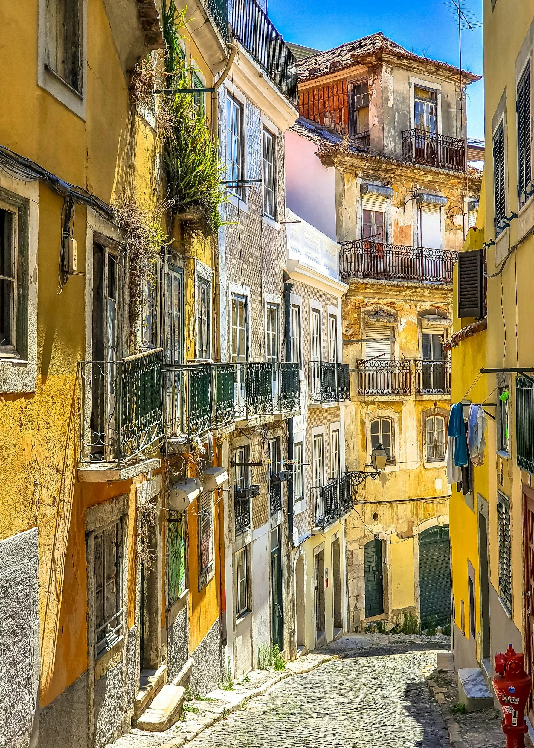 A narrow, cobblestone street in a European city with colorful, weathered buildings, some with balconies and shutters, and laundry hanging outside on the right.