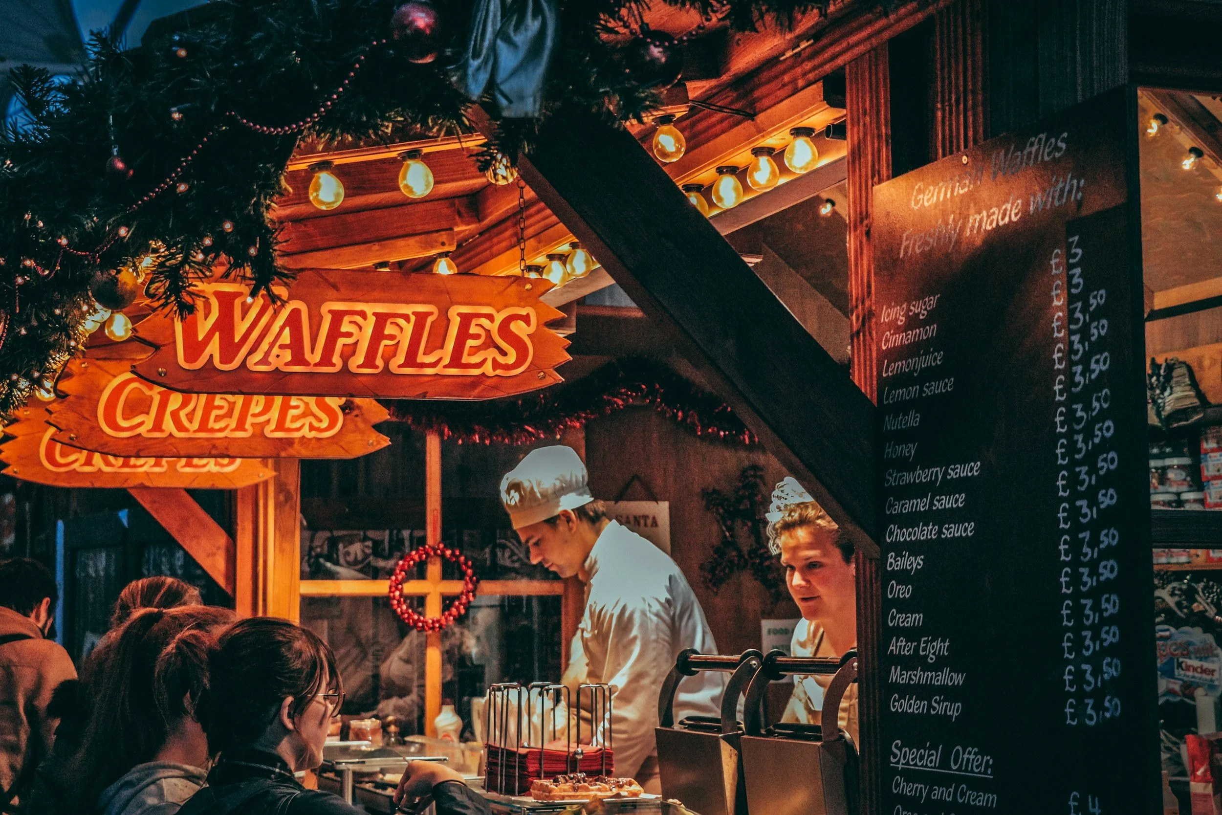 A festive market stall selling waffles, crepes, and various toppings. There are signs for waffles and crepes with customers ordering, and two women behind the counter preparing the food. The stall is decorated with Christmas ornaments and lights.